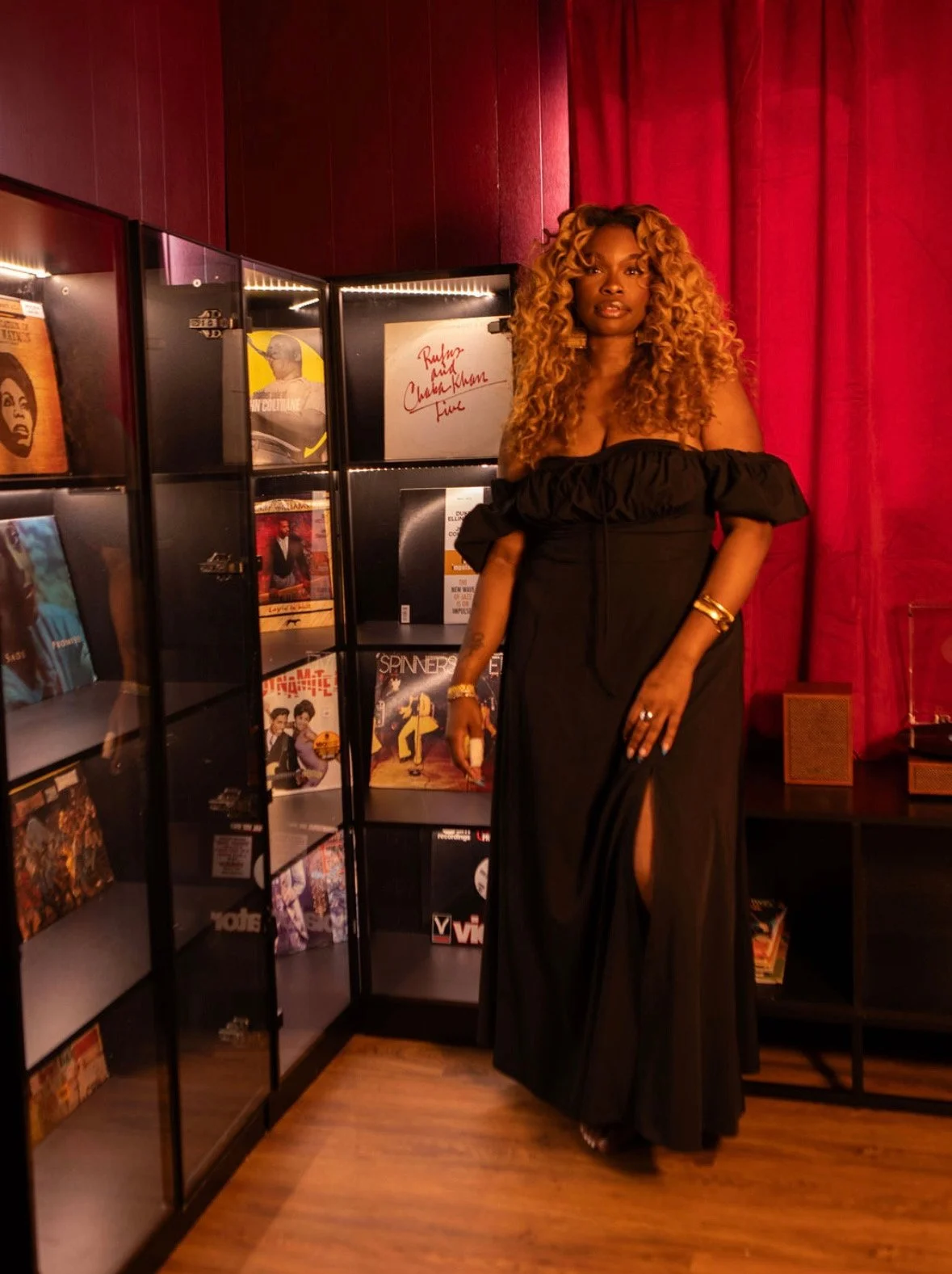 A woman wearing a long black dress with a slit, standing next to a black bookshelf with vinyl records, in a room with red curtains.