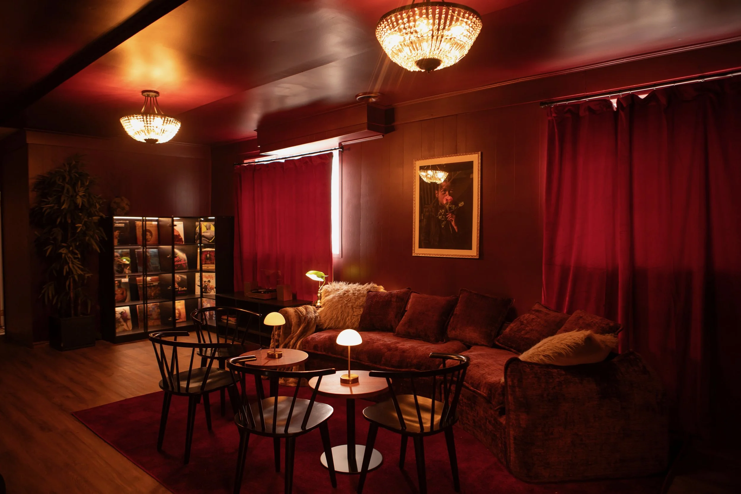 A cozy, dimly lit living room with red curtains, plush furniture, a framed portrait, ceiling chandeliers, and a display shelf with vinyl records.