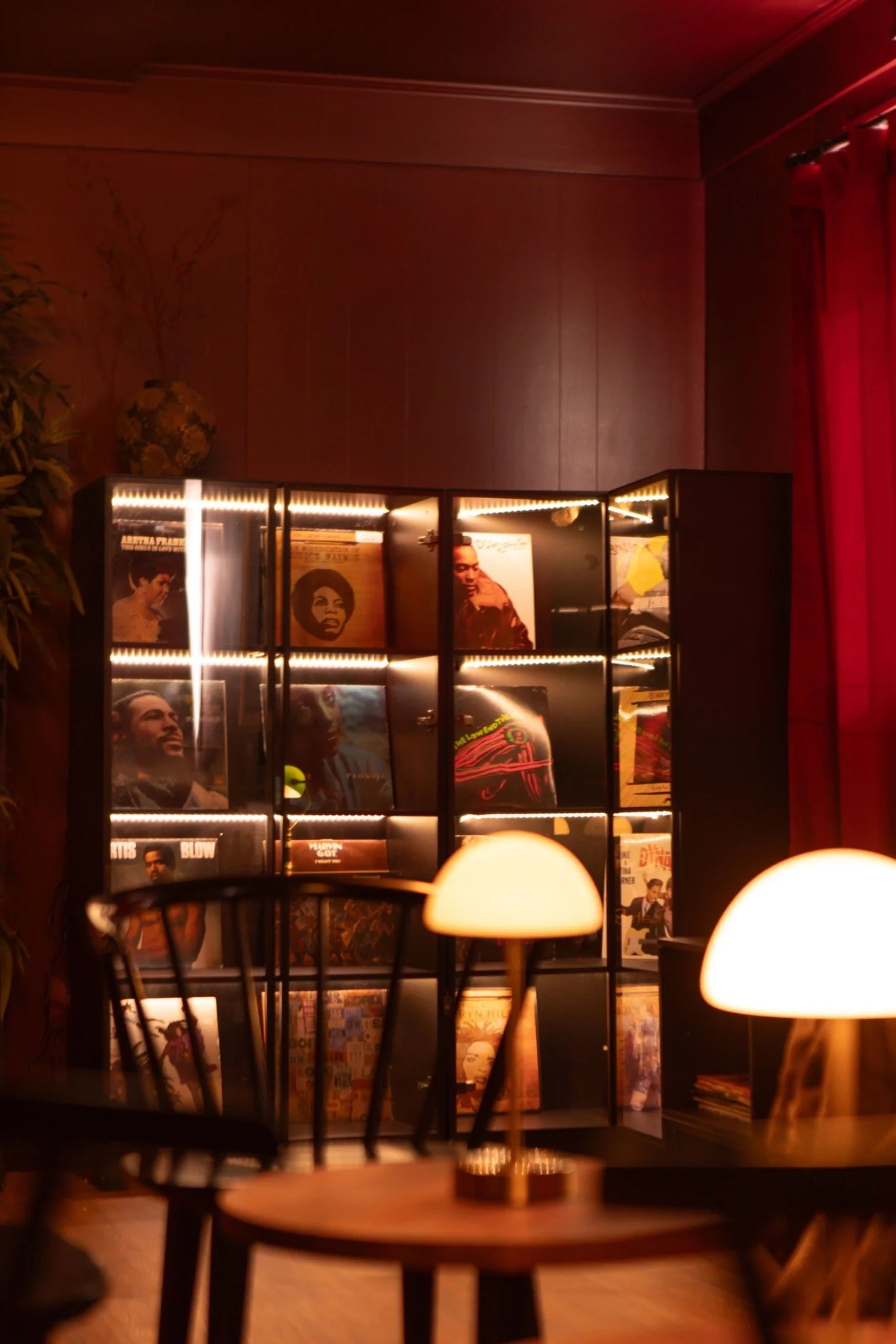 A dimly lit room with a black bookshelf displaying vintage records, surrounded by warm table lamps, red curtains, and dark wooden tables and chairs.