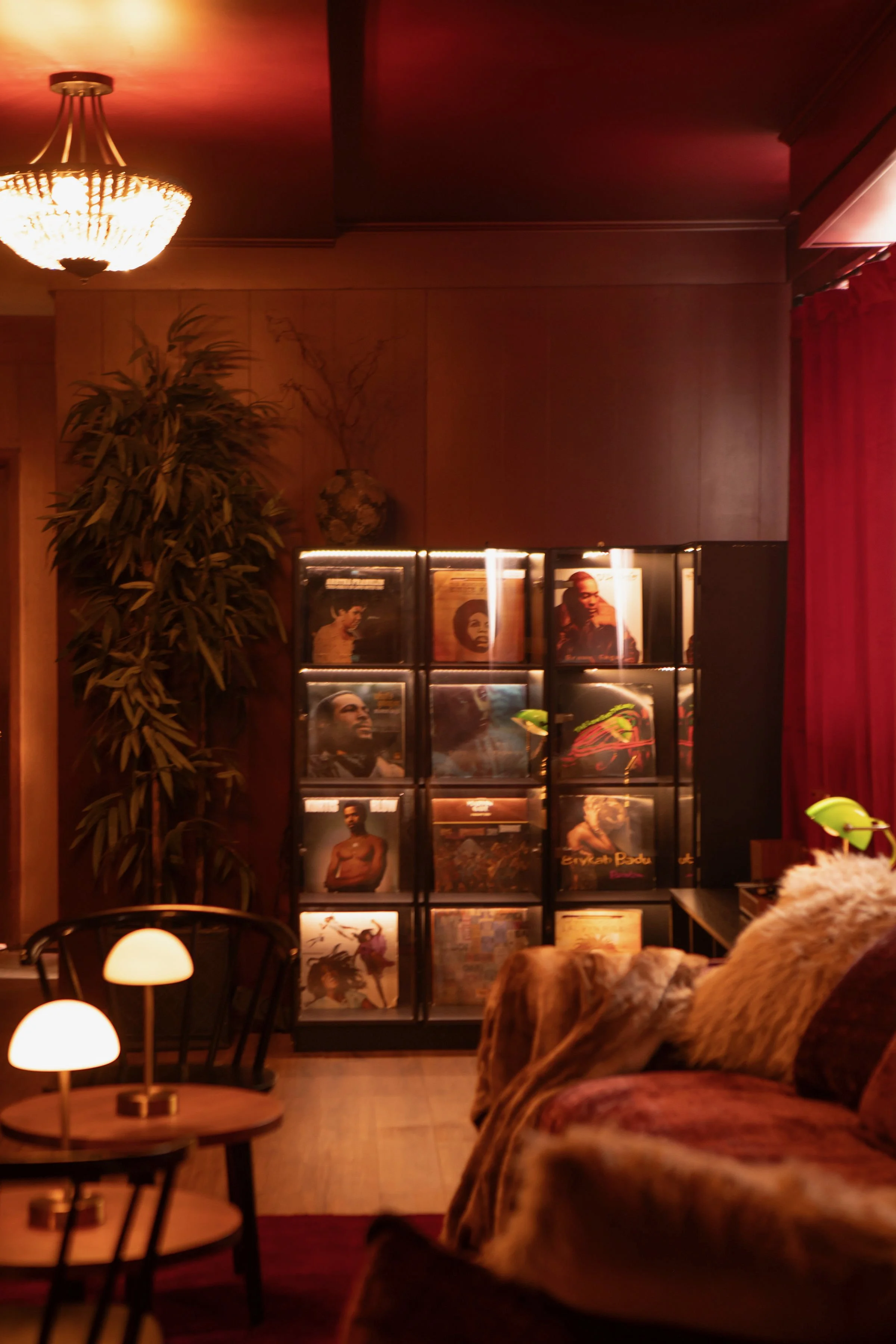 Interior of a room with a wooden wall, a standing bookshelf with magazine covers, a large plant, a small round table with a lamp, and red curtains on the right side.
