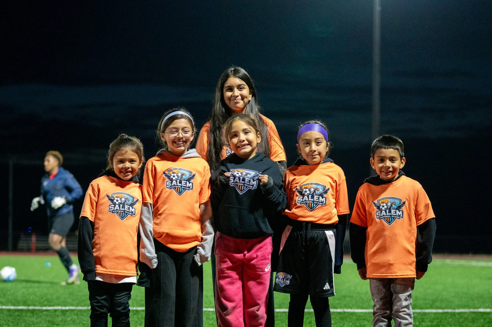 A group of five young children and their female coach stand together on a soccer field at night, all wearing soccer jerseys with the Salem Futbol Club logo. The children are smiling, and the coach is smiling as well. In the background, a young man in a blue jacket is walking across the field.
