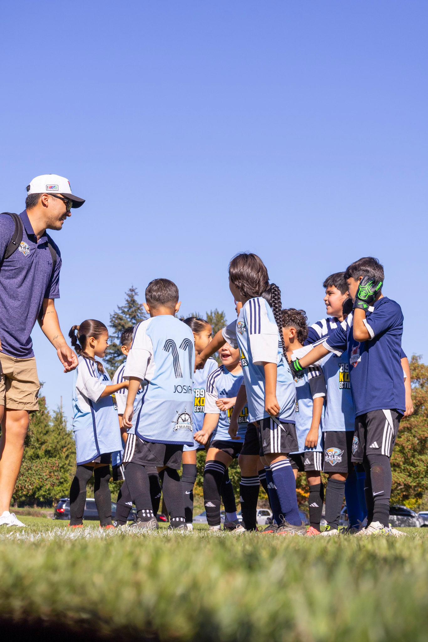 A group of children in soccer uniforms gather for a huddle, with a coach or adult standing to their left during daytime on a soccer field.