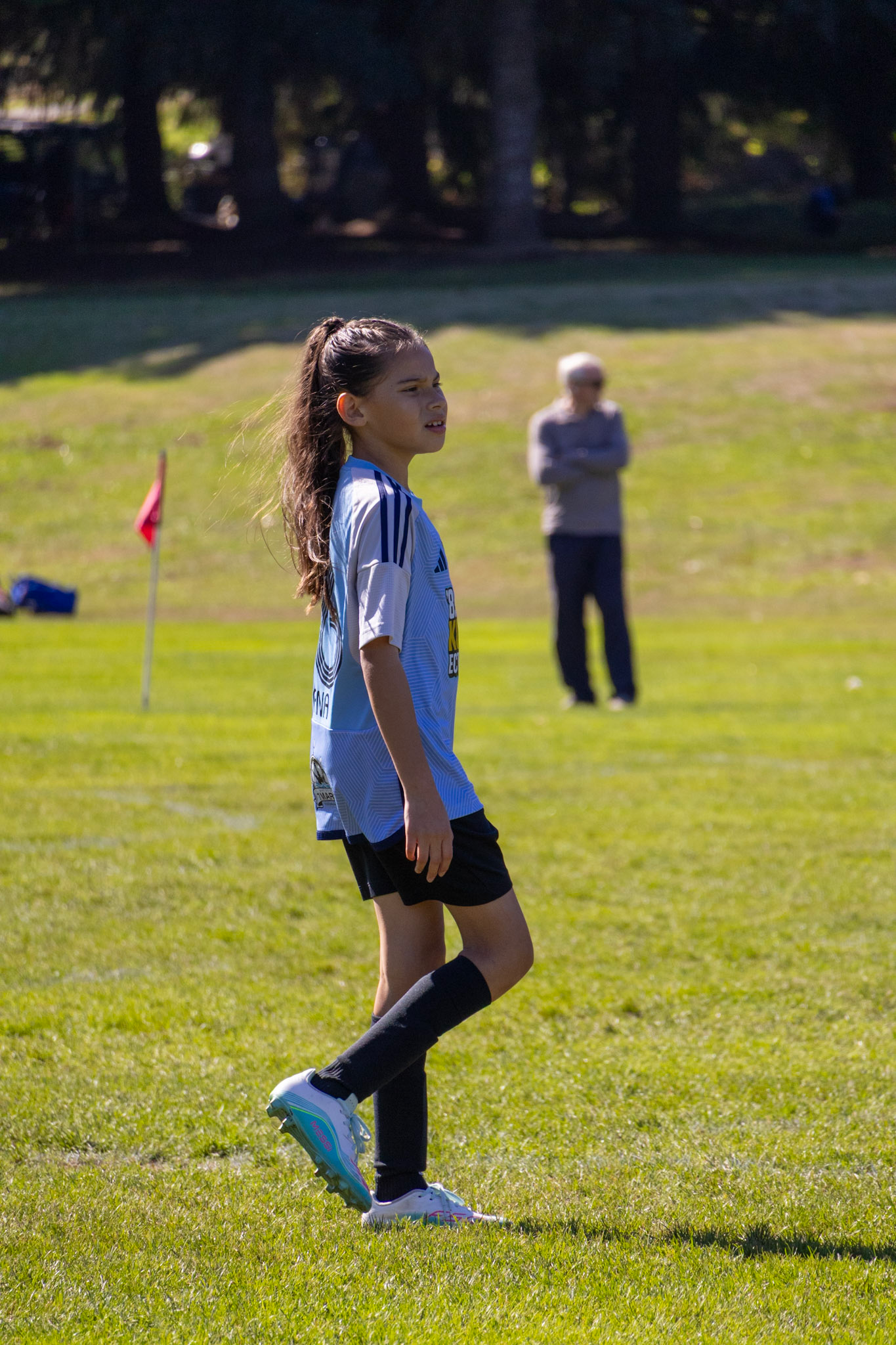 A young girl playing soccer on a grass field with an older woman watching in the background.