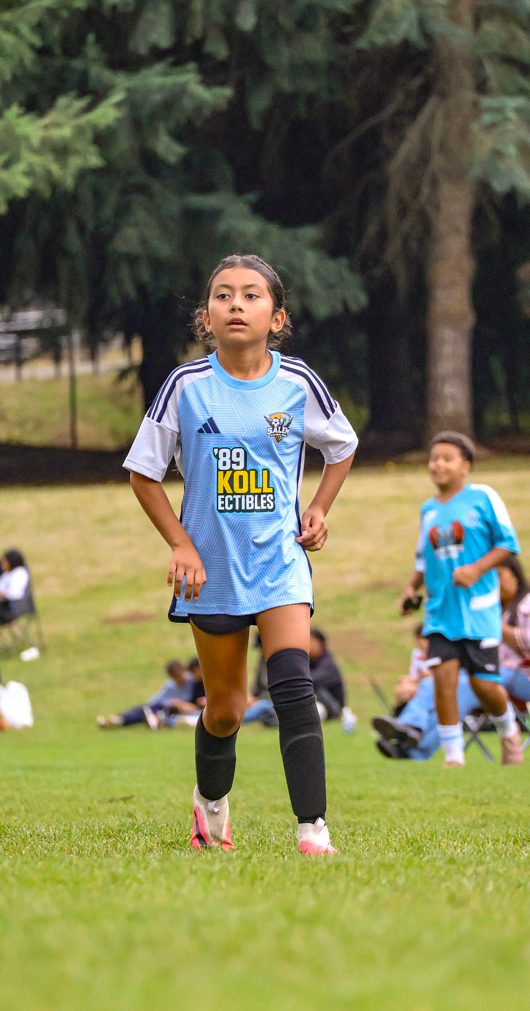 Young girl with dark hair in a braid, wearing a light blue soccer jersey with dark shorts, black knee-high socks, and white athletic shoes, running on a grassy field during a soccer game, with other children and spectators in the background and trees lining the field.