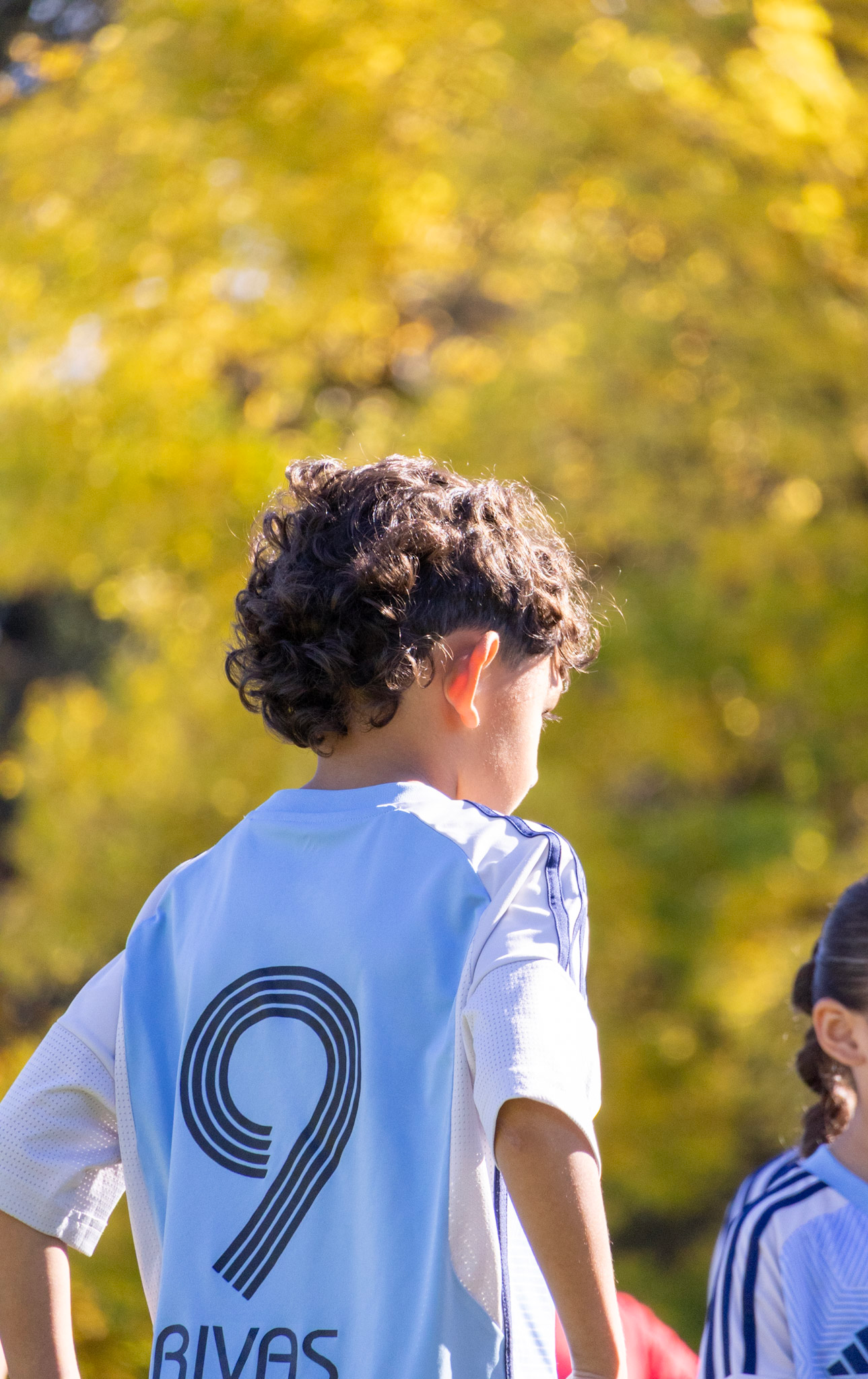 A person with curly hair wearing a white sports jersey with black text and a circular logo on the back, standing outdoors with trees in the background.