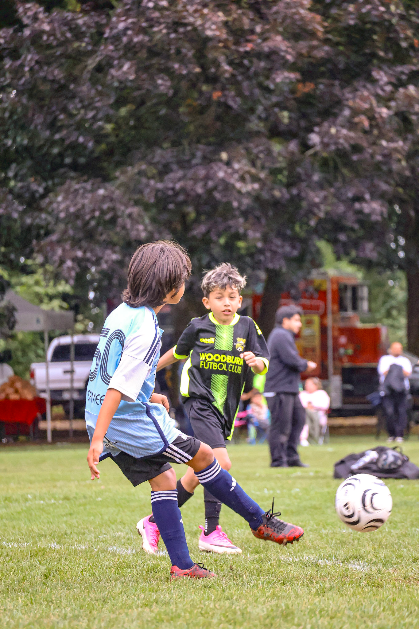Two young boys playing soccer on a grassy field, with a large tree and people in the background. One boy is wearing a light blue soccer uniform, and the other is in a black and green jersey that says "Woodbury Futbol Club." Both are focused on the soccer ball.