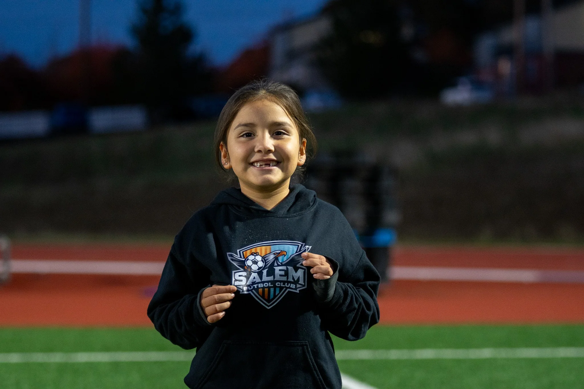 A young girl smiling and holding a soccer team logo sticker on her black hoodie, standing on an outdoor sports field during dusk.