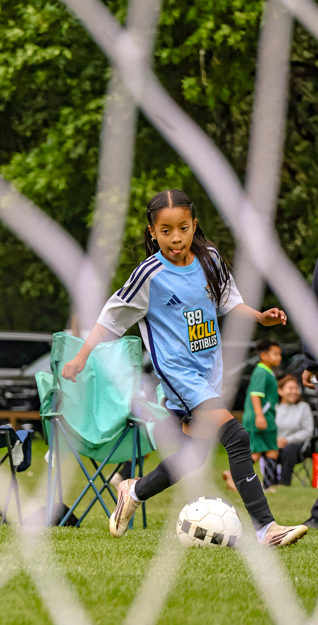 Young girl in a blue sports jersey kicking a soccer ball during a game, viewed through a chain-link fence with a grassy field background.