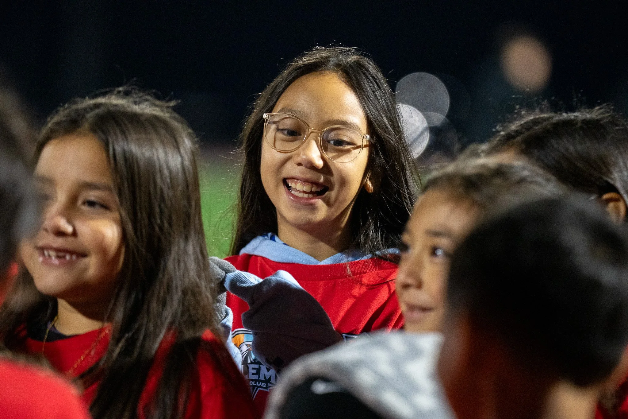 A group of children smiling and gathered closely together outdoors at night, with bright lights and a dark sky in the background.