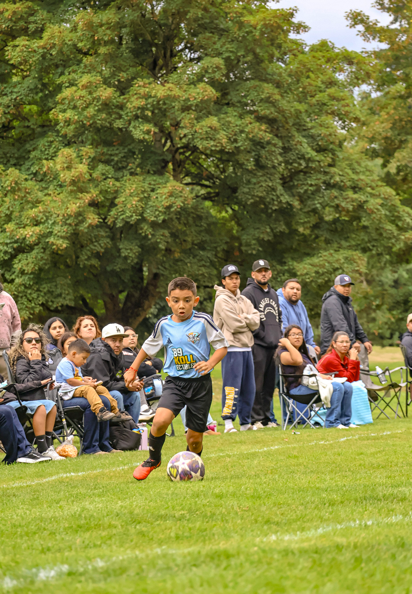 A young boy playing soccer on a grass field, with a crowd of spectators sitting and standing behind him. The background features large, green trees.
