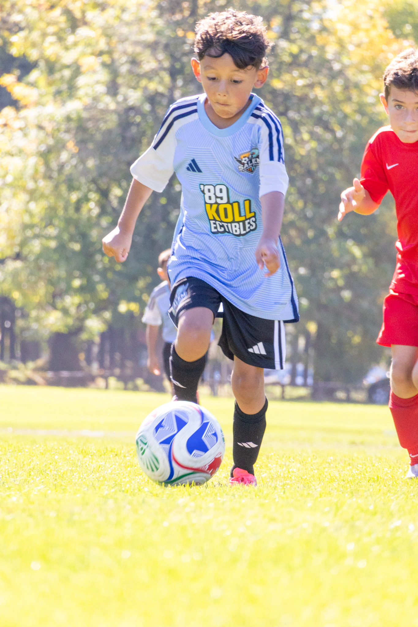 Two young boys playing soccer on a grassy field during daytime, with one boy in a white and blue jersey and the other in a red jersey.