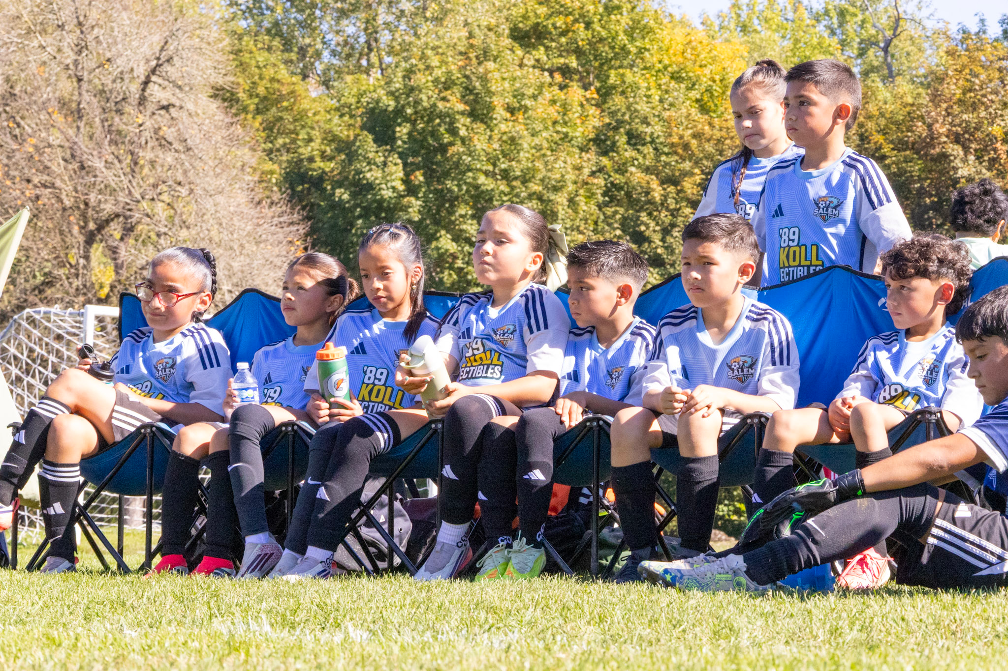 Children sitting on a bench watching a soccer game outdoors on a sunny day, with trees in the background.