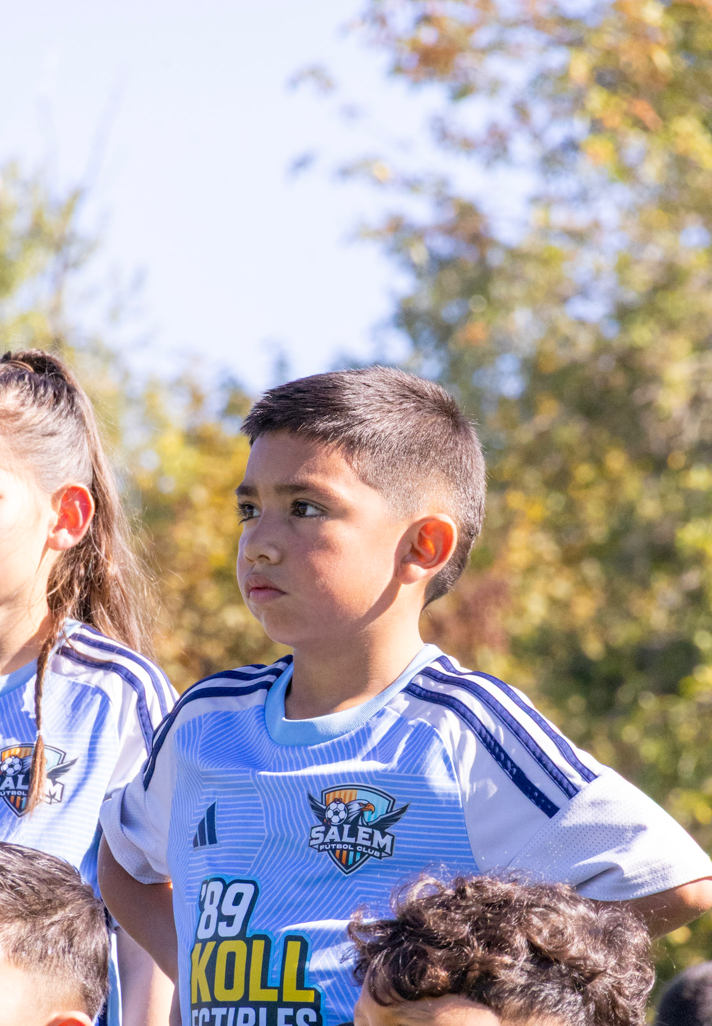 Close-up of a young boy with short dark hair, wearing a white and blue soccer jersey with logos, standing outdoors with trees and a blue sky in the background.