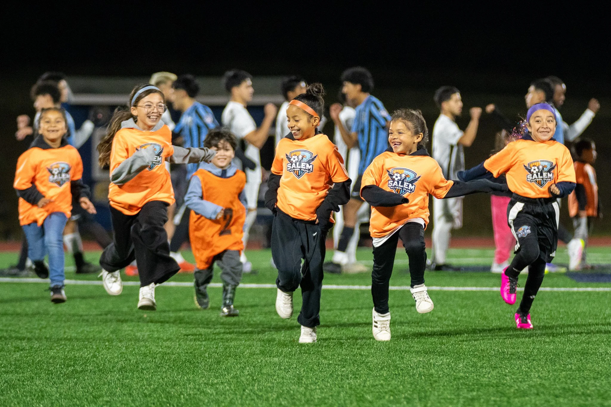 Children running and playing soccer on a field at night, wearing orange and blue sports jerseys with a 'Salem' team logo, smiling and having fun.