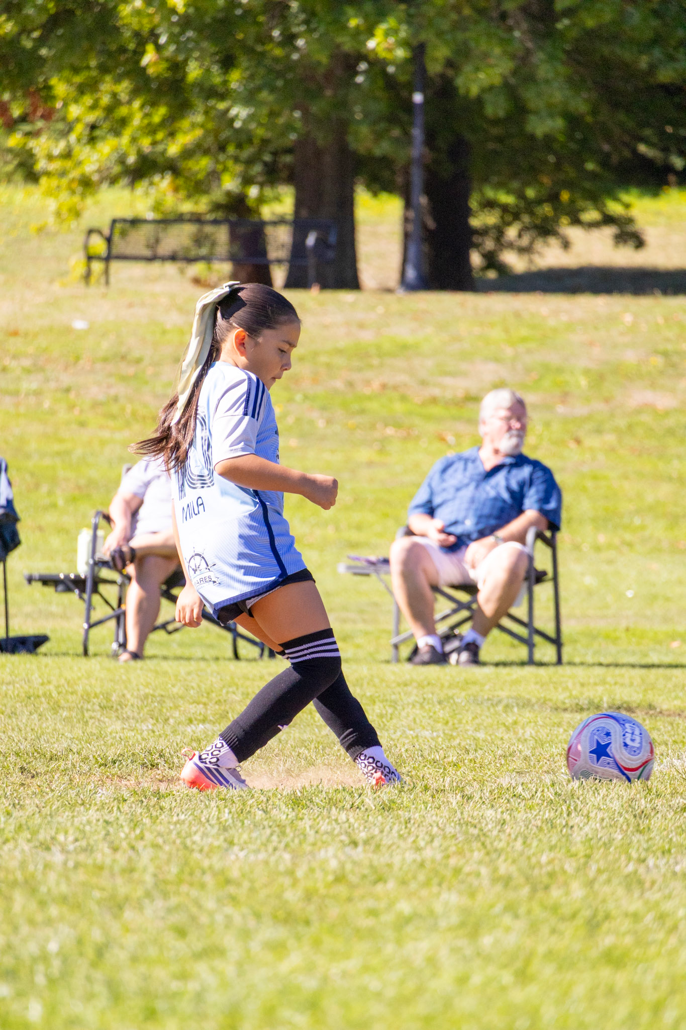 A young girl playing soccer on a grassy field with a soccer ball. She is wearing a white and blue sports uniform, black knee-high socks, and colorful cleats. In the background, there are two seated adults, one with white hair and a beard, watching her play, and there are trees and a park bench.