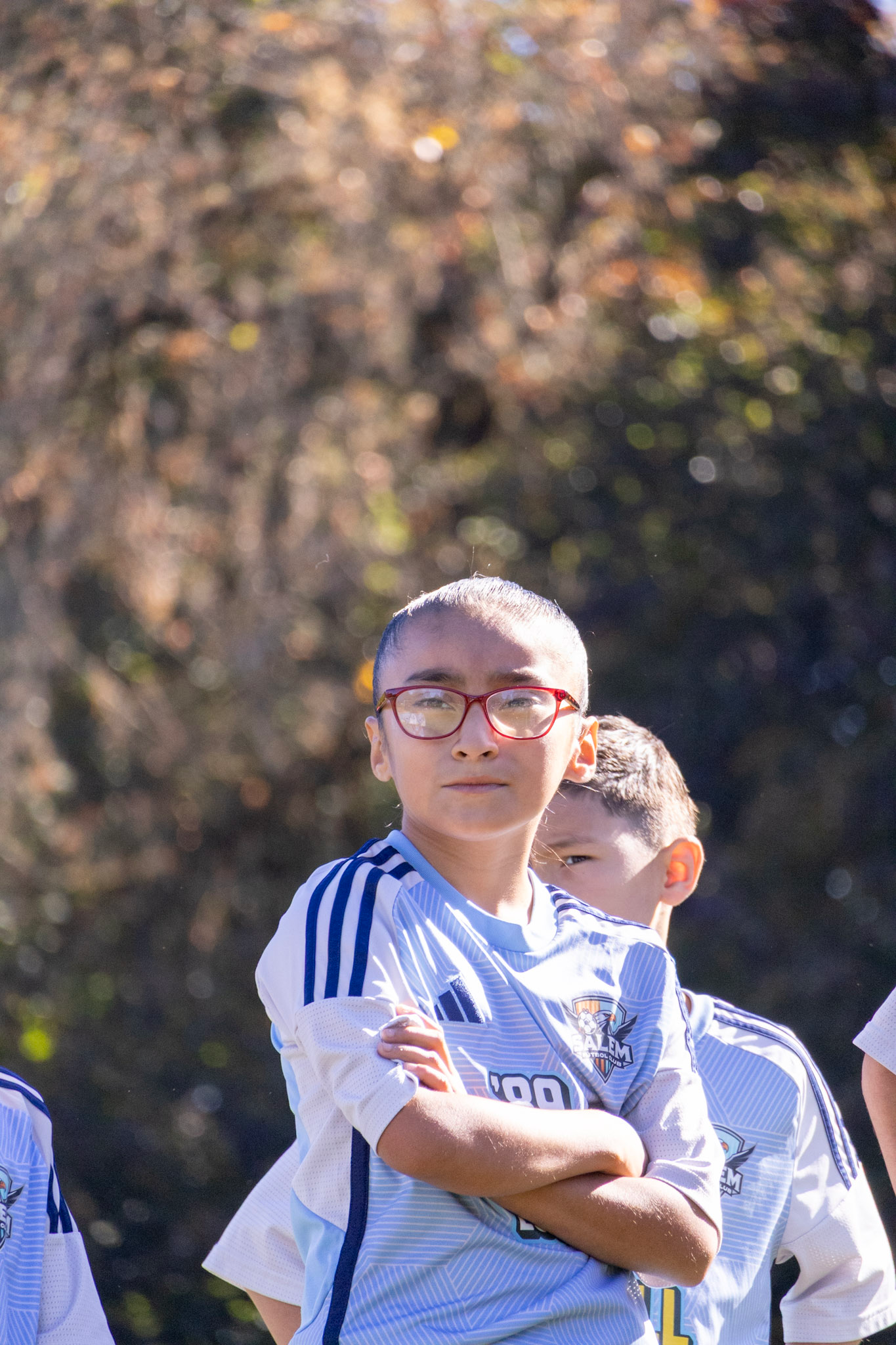 A young boy with glasses and a soccer jersey standing outdoors with his arms crossed.