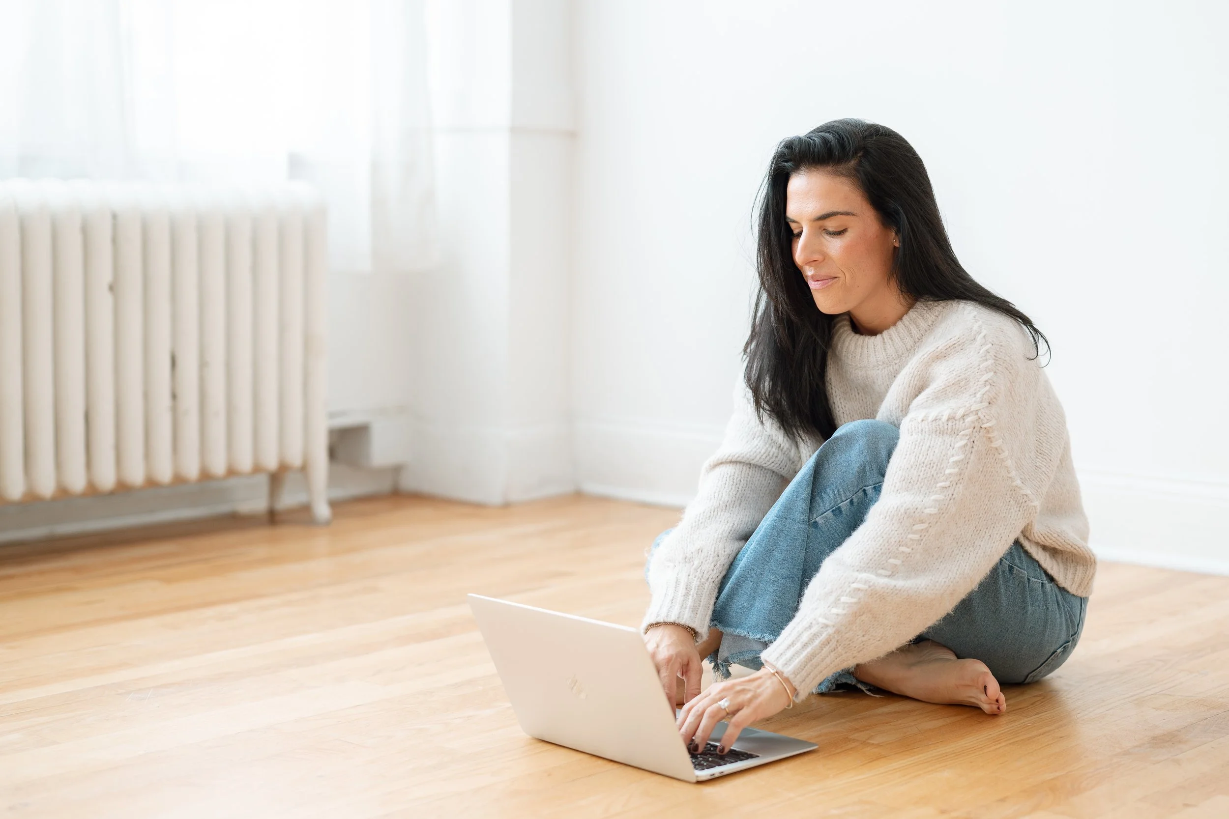 A woman with long black hair sitting on the wooden floor with her legs crossed. She is wearing a beige knit sweater and blue jeans, looking at a laptop in front of her in a bright room with white walls and a large window.