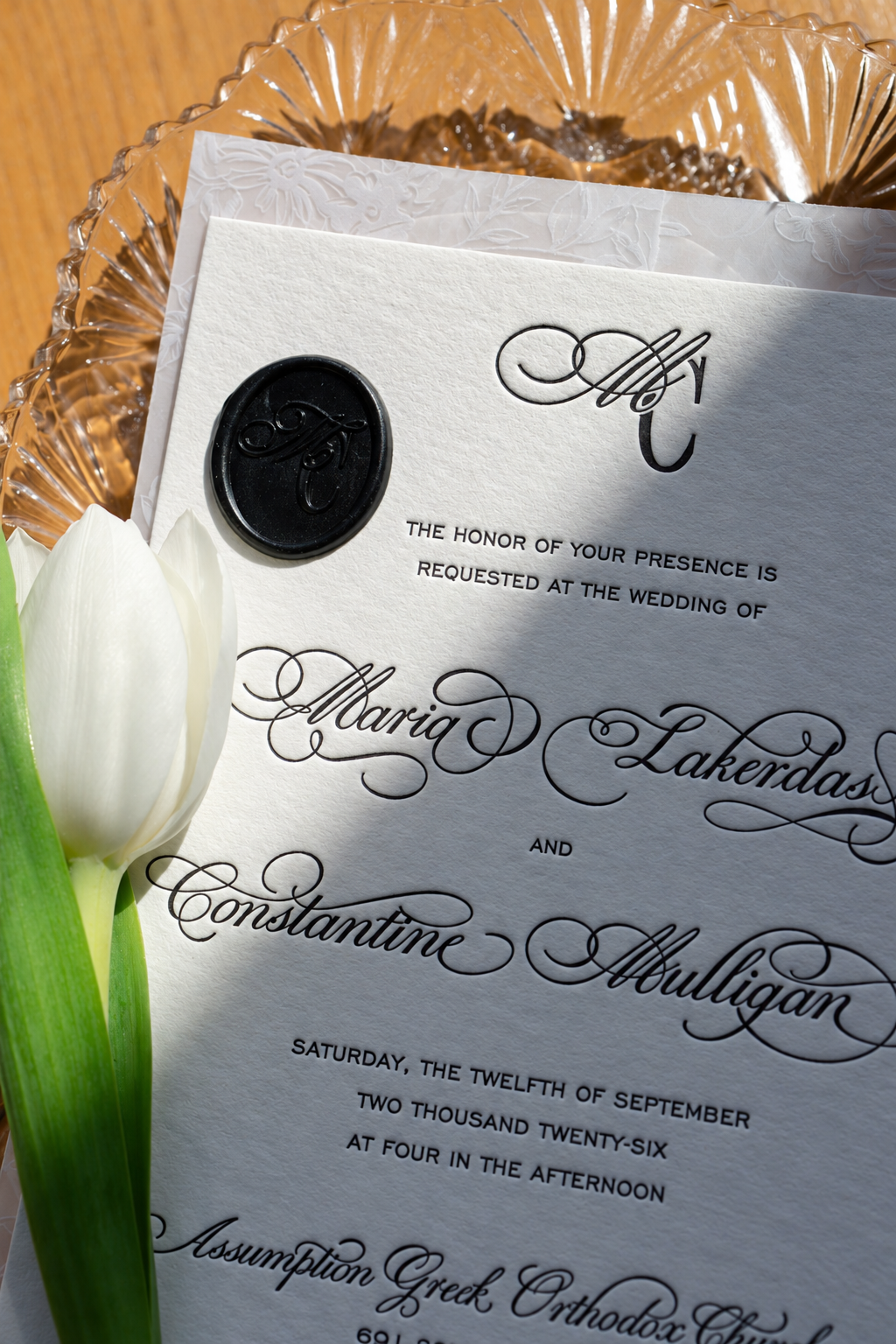 Wedding invitation with black wax seal, featuring elaborate black lettering, placed on a floral background with a white tulip nearby.