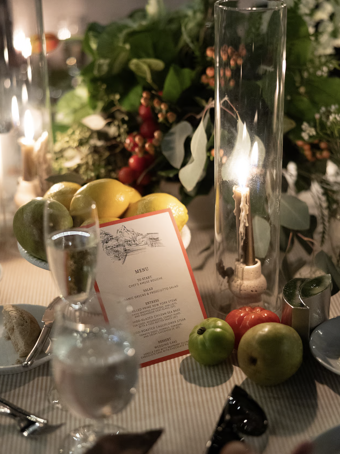 A table setting with lemons, green apple, tomato, and a menu surrounded by greenery and candles.