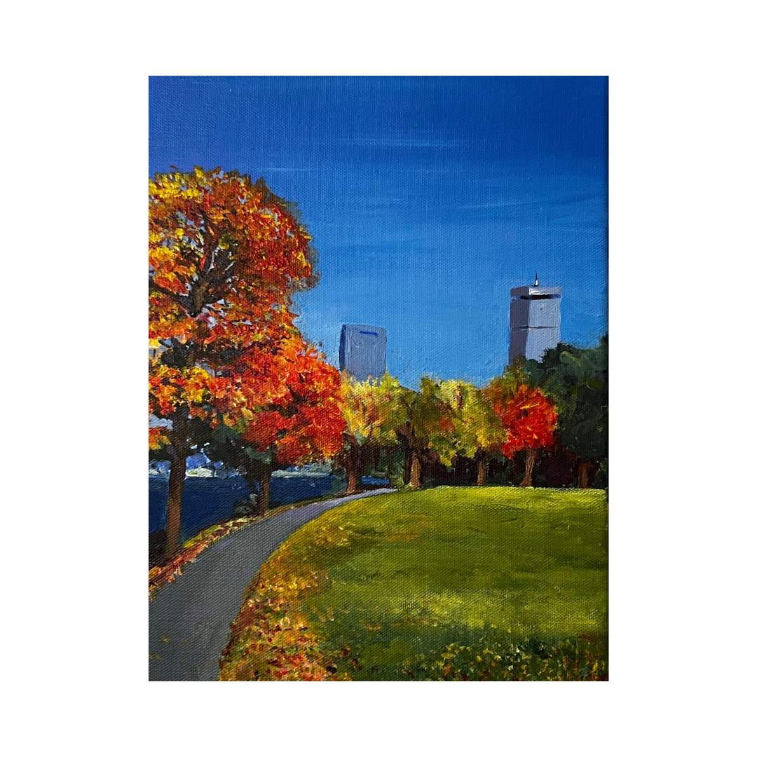 A Boston park by the Charles River with a curved pathway, colorful autumn trees, and skyscrapers in the background under a clear blue sky.