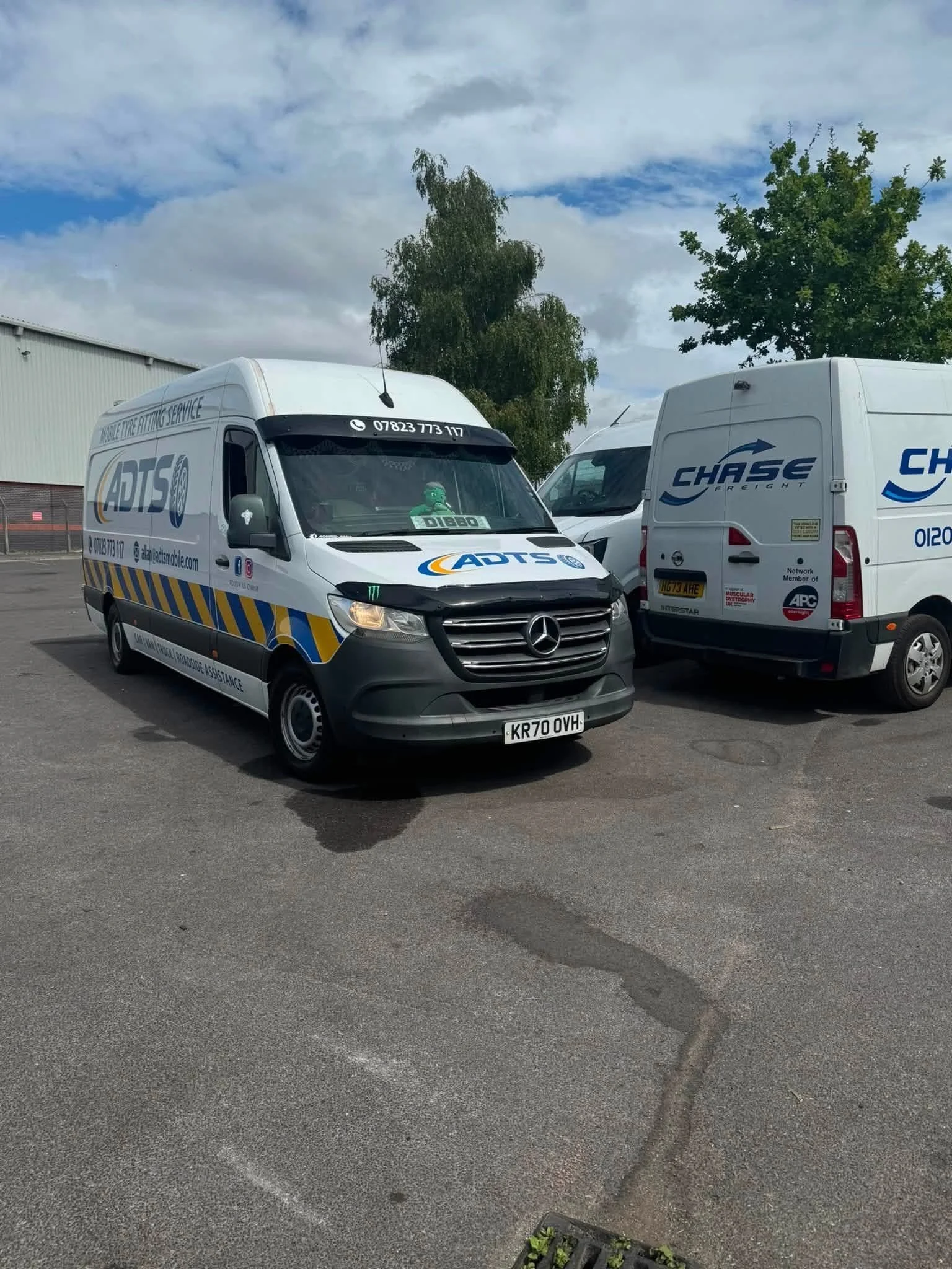 Parking lot with two white delivery vans, one labeled 'ADTS' and the other 'CHASE'. There are trees and a building with a corrugated metal exterior in the background, under a partly cloudy sky.