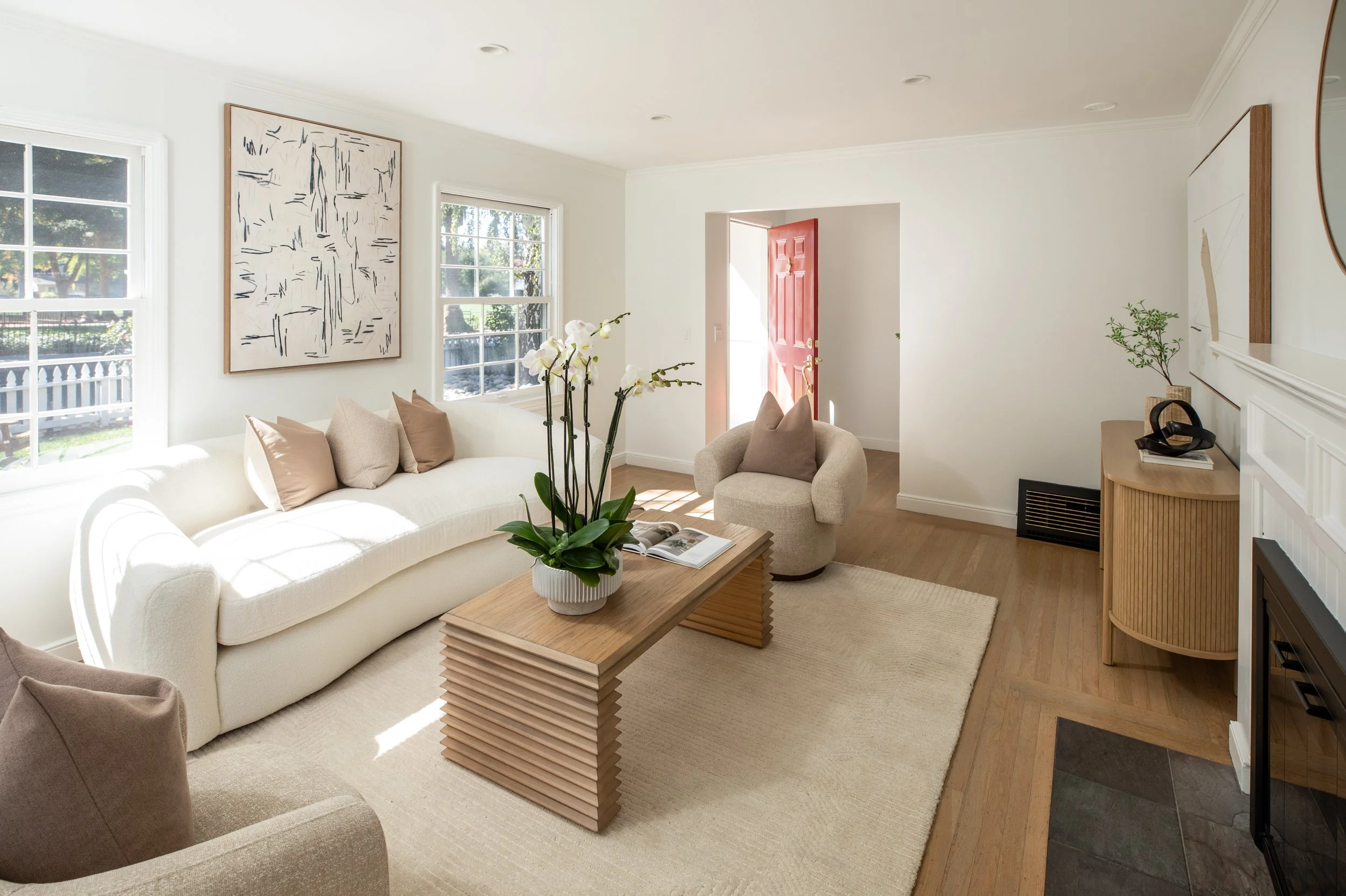 A living room with a white couch, beige pillows, a wooden coffee table with a plant, and a fireplace, with windows letting in sunlight.