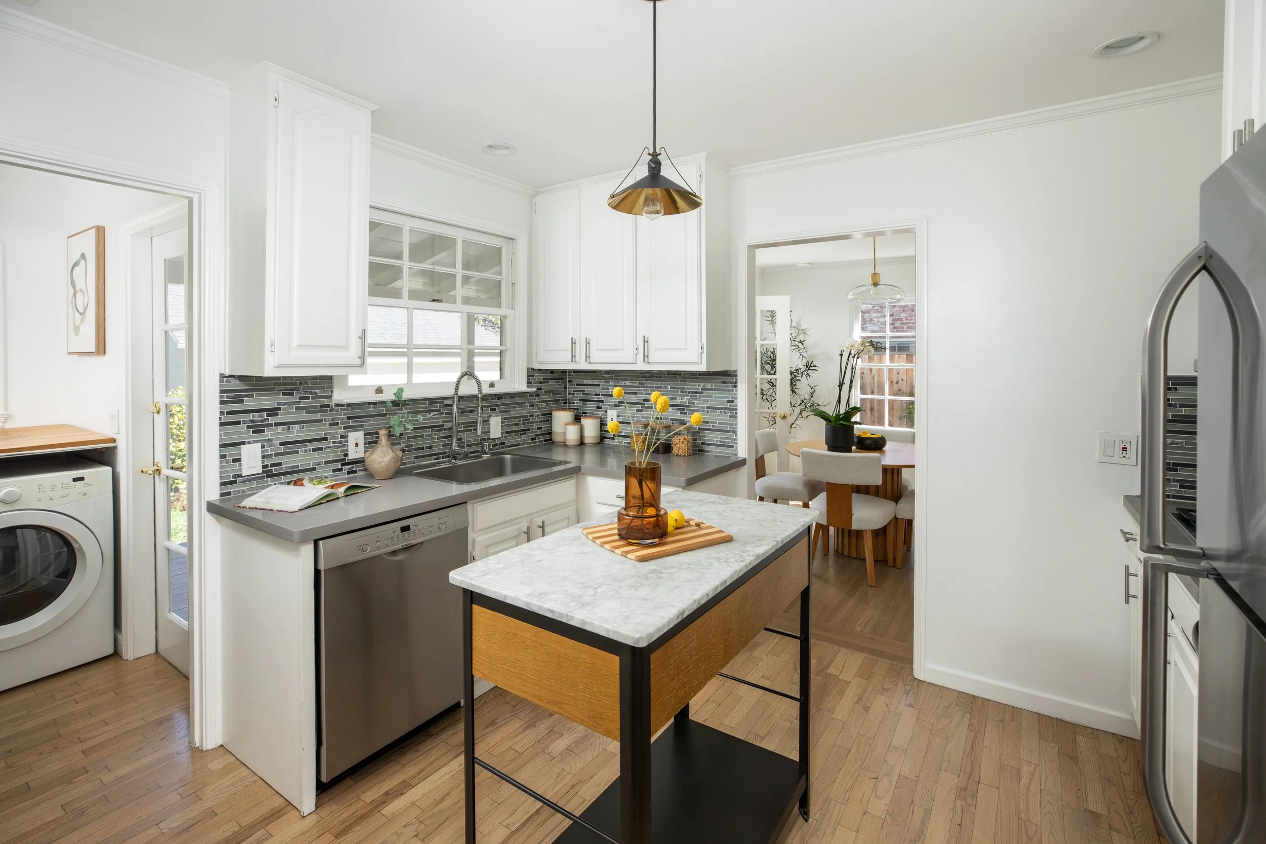 A modern kitchen with white cabinets, gray countertops, a gray tile backsplash, and a small kitchen island with a marble top. There are yellow flowers in a glass vase on the island, and a window behind the sink. Adjacent to the kitchen is a dining ar