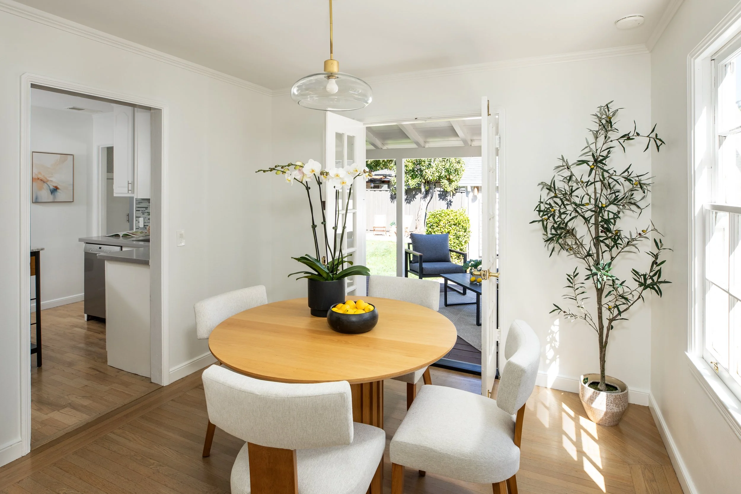 A cozy dining area with a round wooden table surrounded by white chairs, a vase of white orchids, and a bowl of lemons. The space opens onto an outdoor patio with a blue chair and greenery visible outside.