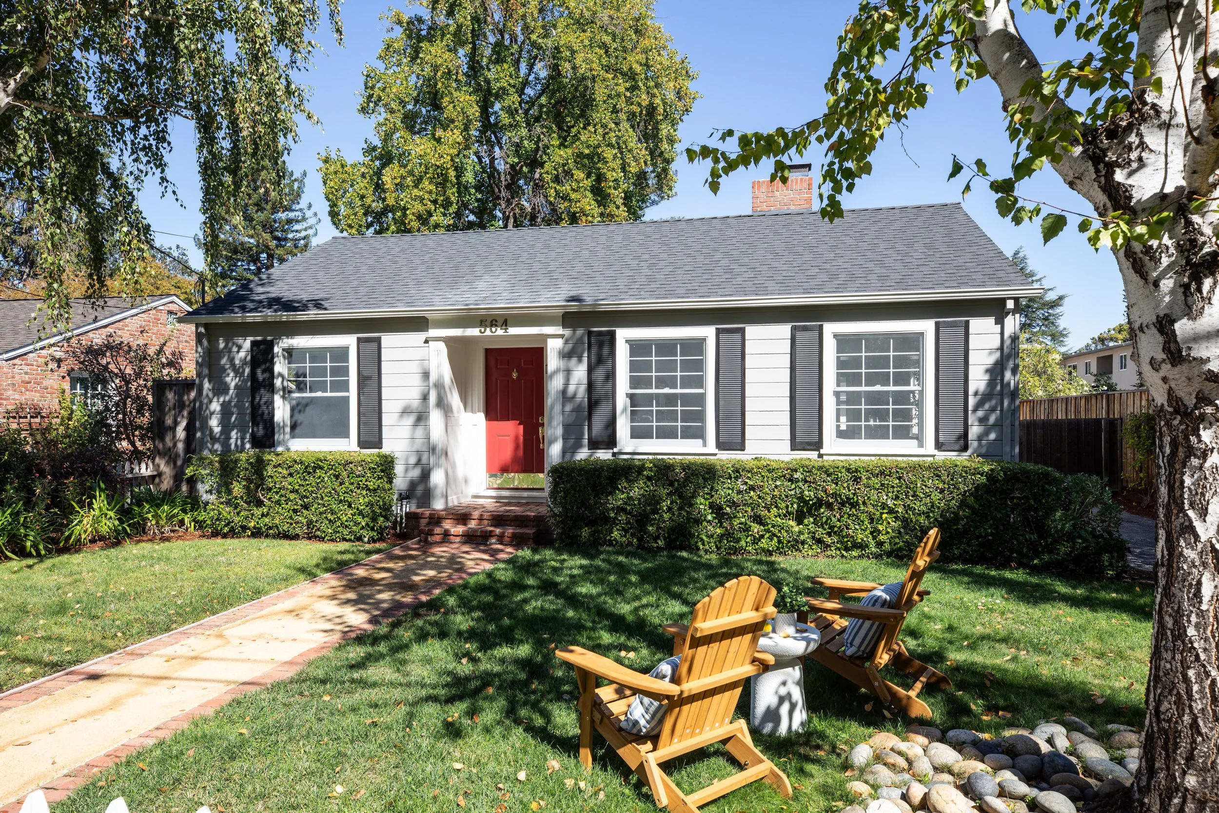 Front view of a white house with black shutters and a red door, surrounded by lush greenery and trees, with a paved walkway leading to the entrance, and outdoor seating in the yard.