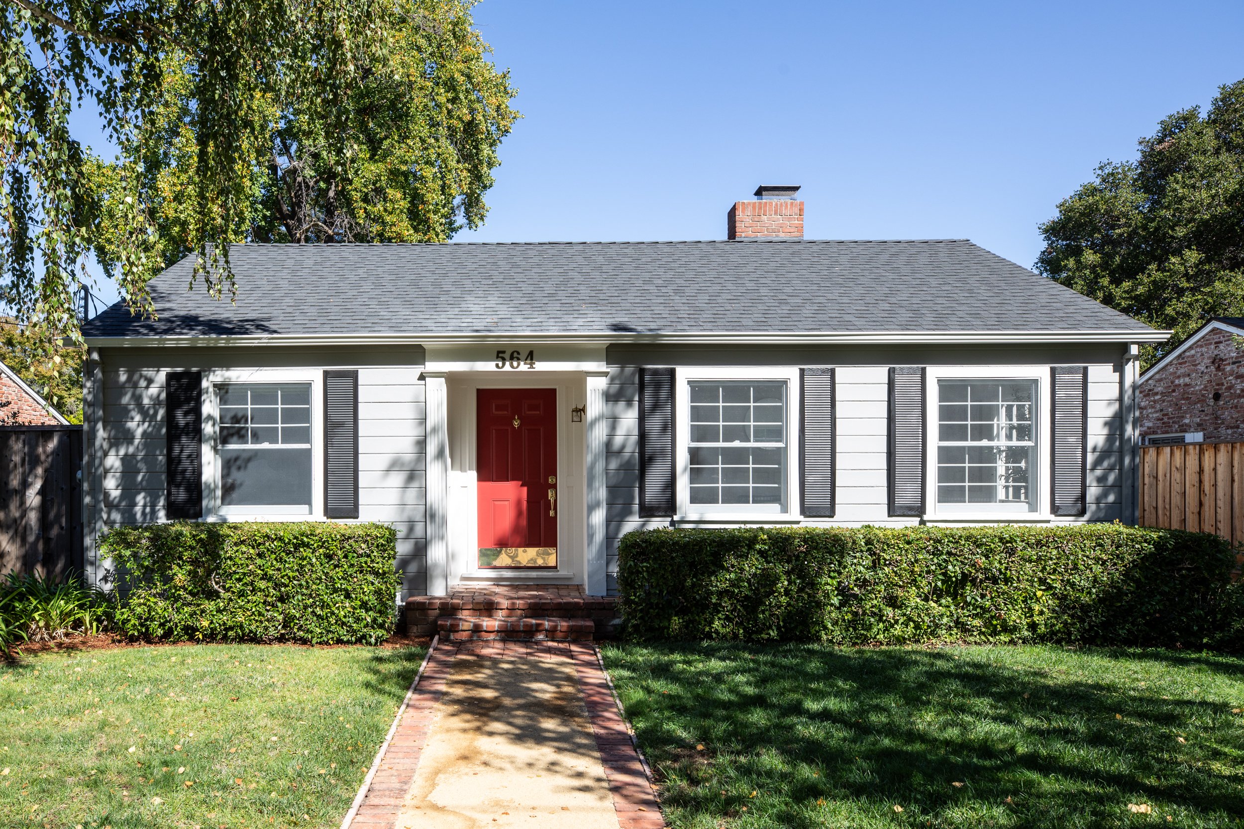 Front view of a house with white siding, a red front door, black shutters, a brick chimney, and a brick walkway leading to the entrance. The house has a well-maintained lawn and surrounding trees.