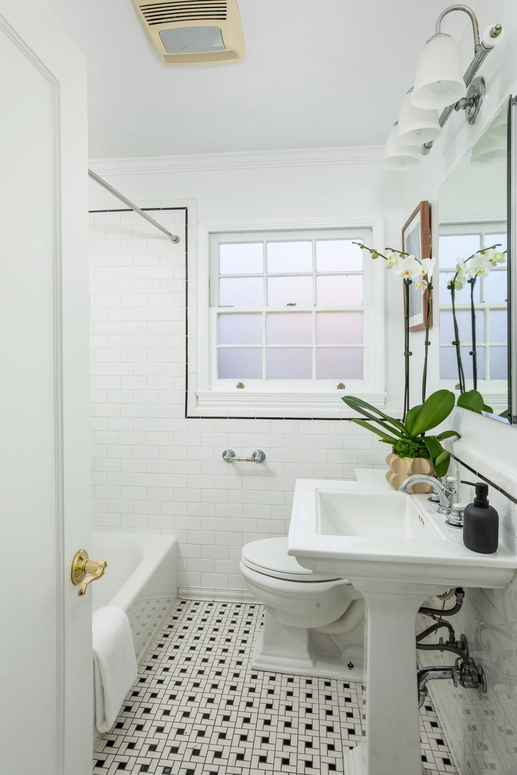 A small vintage-style bathroom with a white pedestal sink, a toilet, and a window with frosted glass. The walls are white with white subway tiles around the shower area, which has a black border. There is a potted orchid on the sink, and a mirror wit