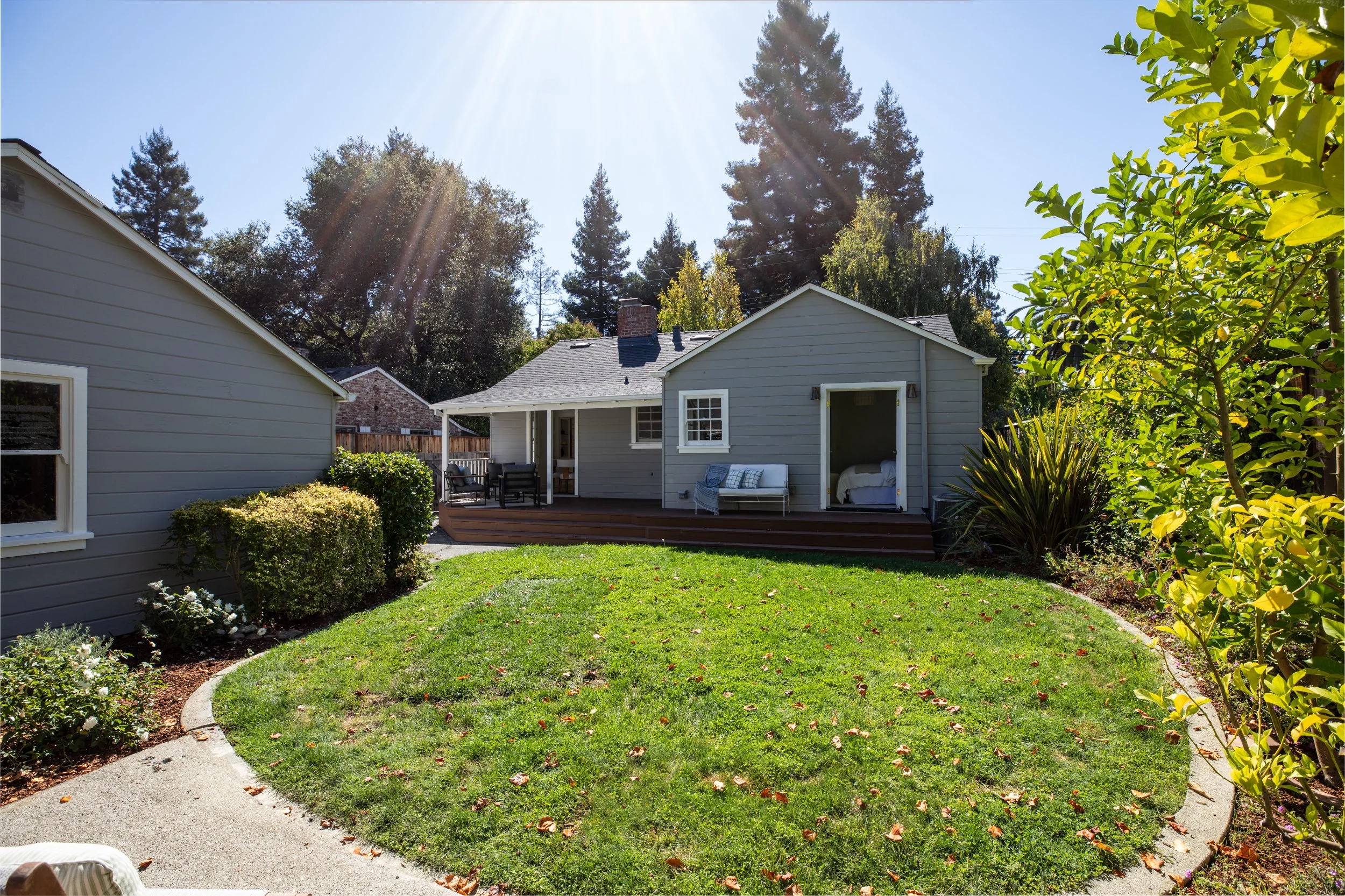 A backyard with a well-maintained lawn, a concrete pathway, flower beds, and a gray house with a porch and outdoor furniture, with tall trees and a clear sunny sky in the background.