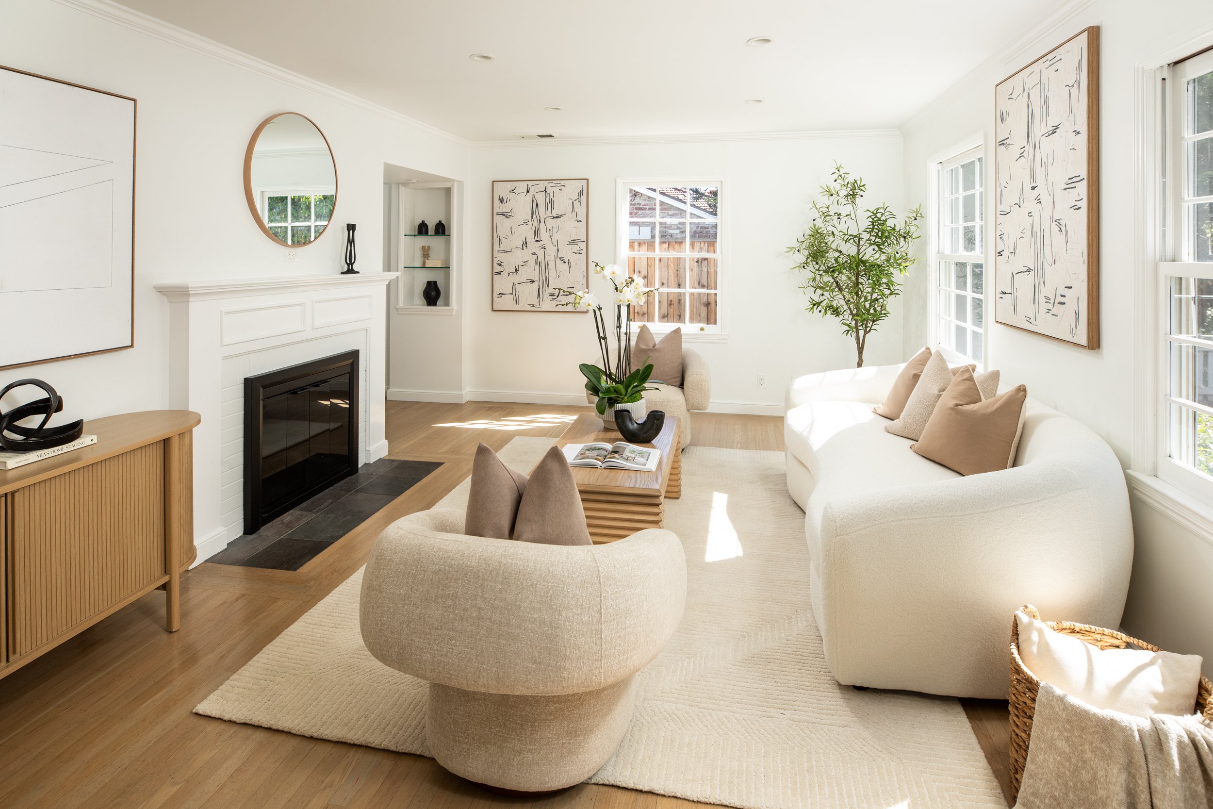Bright living room with white walls, large windows, a white sofa with beige pillows, a round beige armchair, a wooden coffee table with a plant, artwork on the walls, a fireplace, and natural light