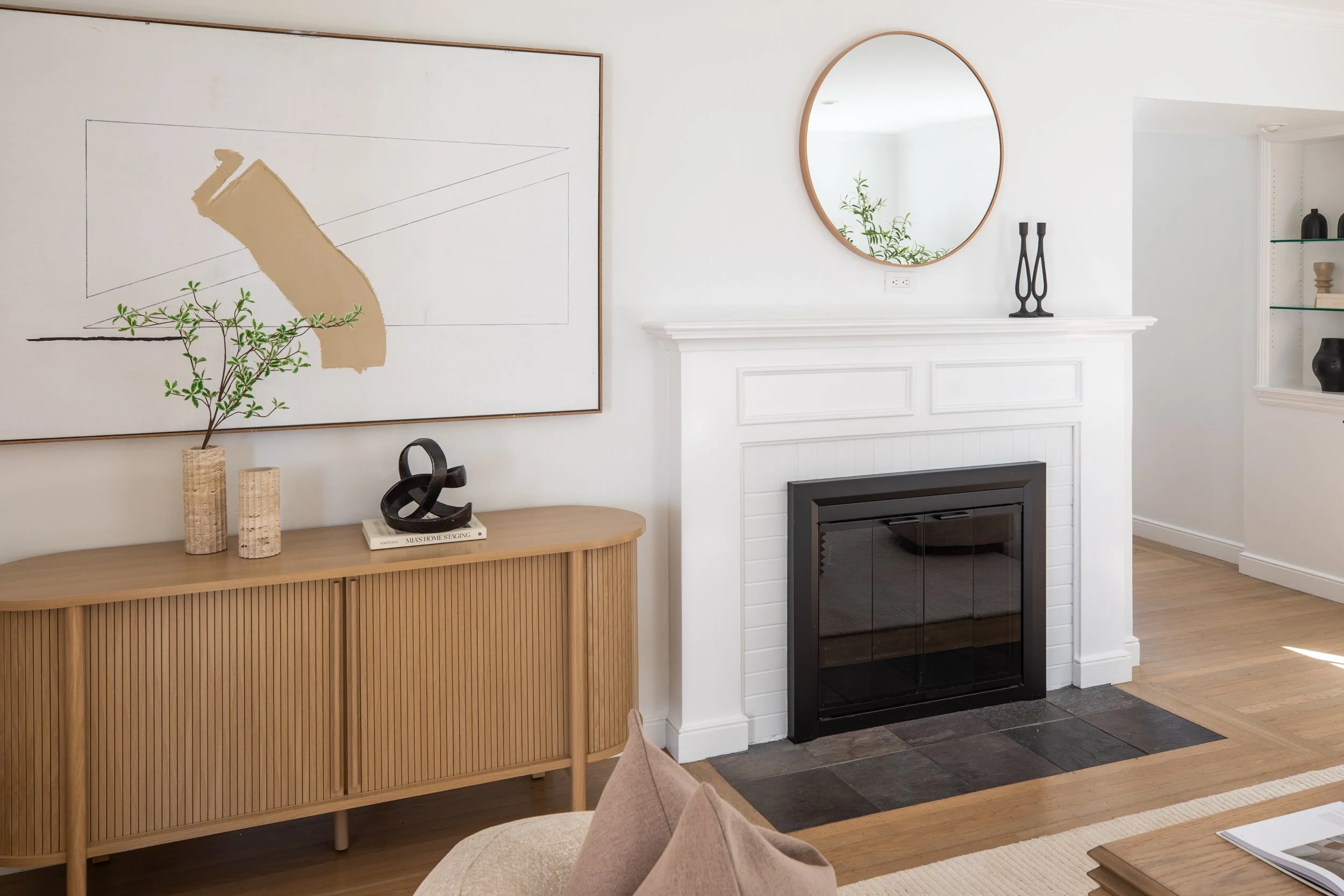 Interior of a living room with a white fireplace, a round mirror, modern decorative objects, and a wooden sideboard with vases and a book, in a minimalist style.