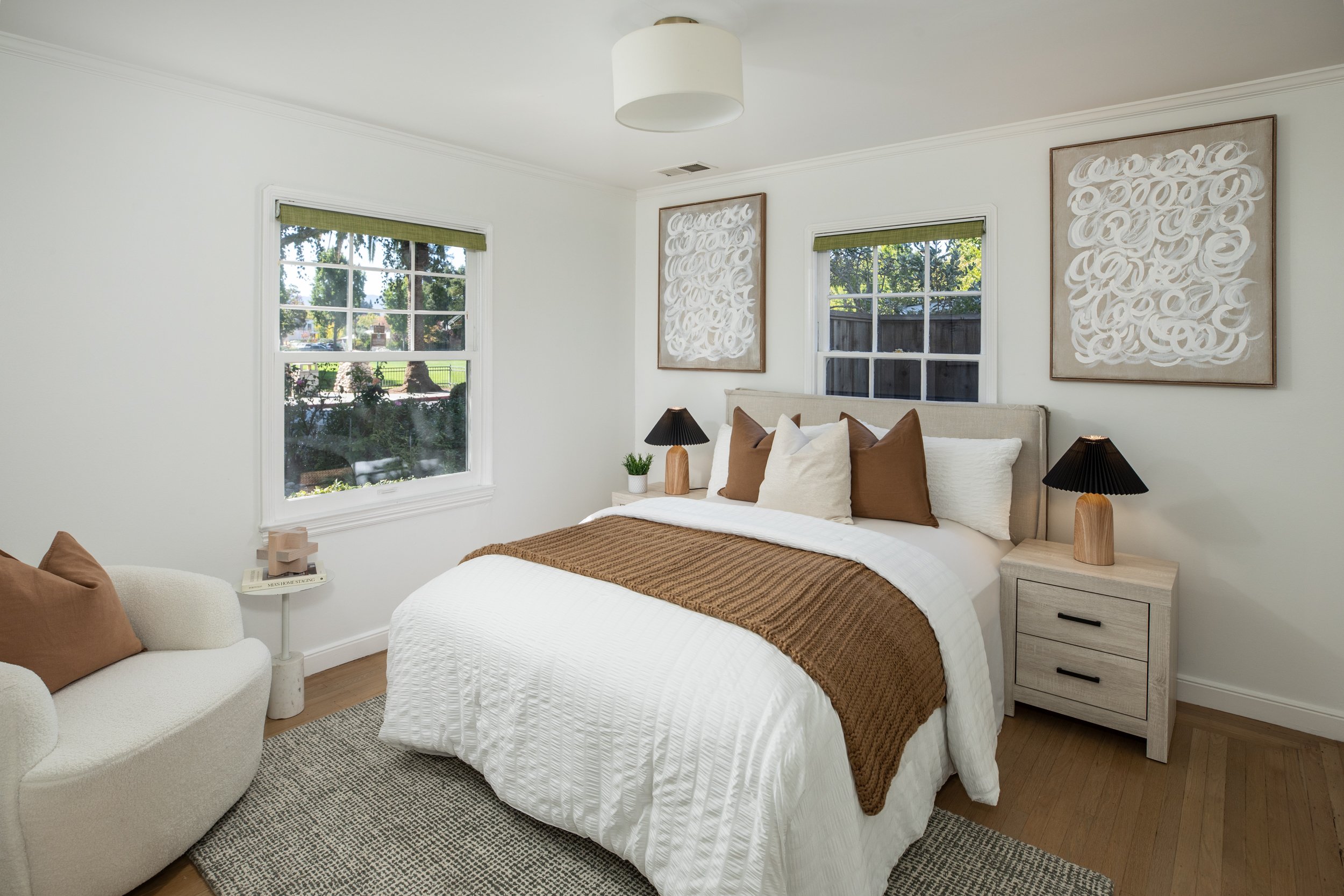 A bedroom with a bed, pillows, and a blanket. There are two nightstands with lamps, a small table with a plant, and a white armchair with a brown pillow. Artwork hangs above the bed, and windows show a sunny outdoor view with trees and a fence.