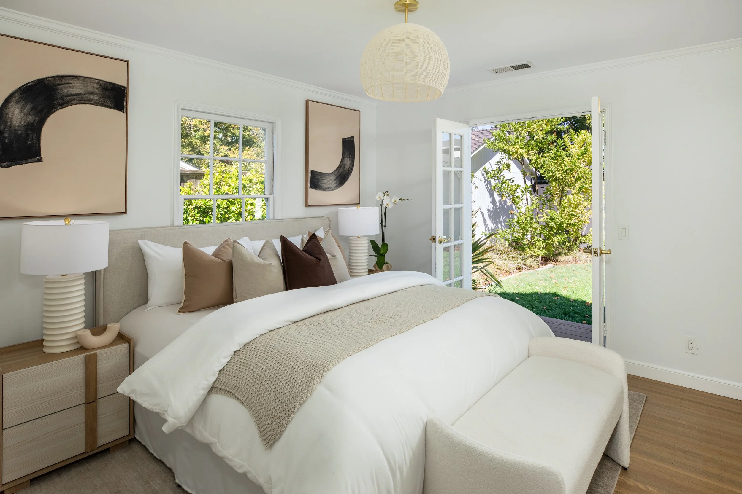 A cozy bedroom with a large bed, beige and white bedding, throw pillows, and a small plant beside the bed. There are abstract art paintings on the white walls, and natural light streaming in through open French doors and windows with views of greener