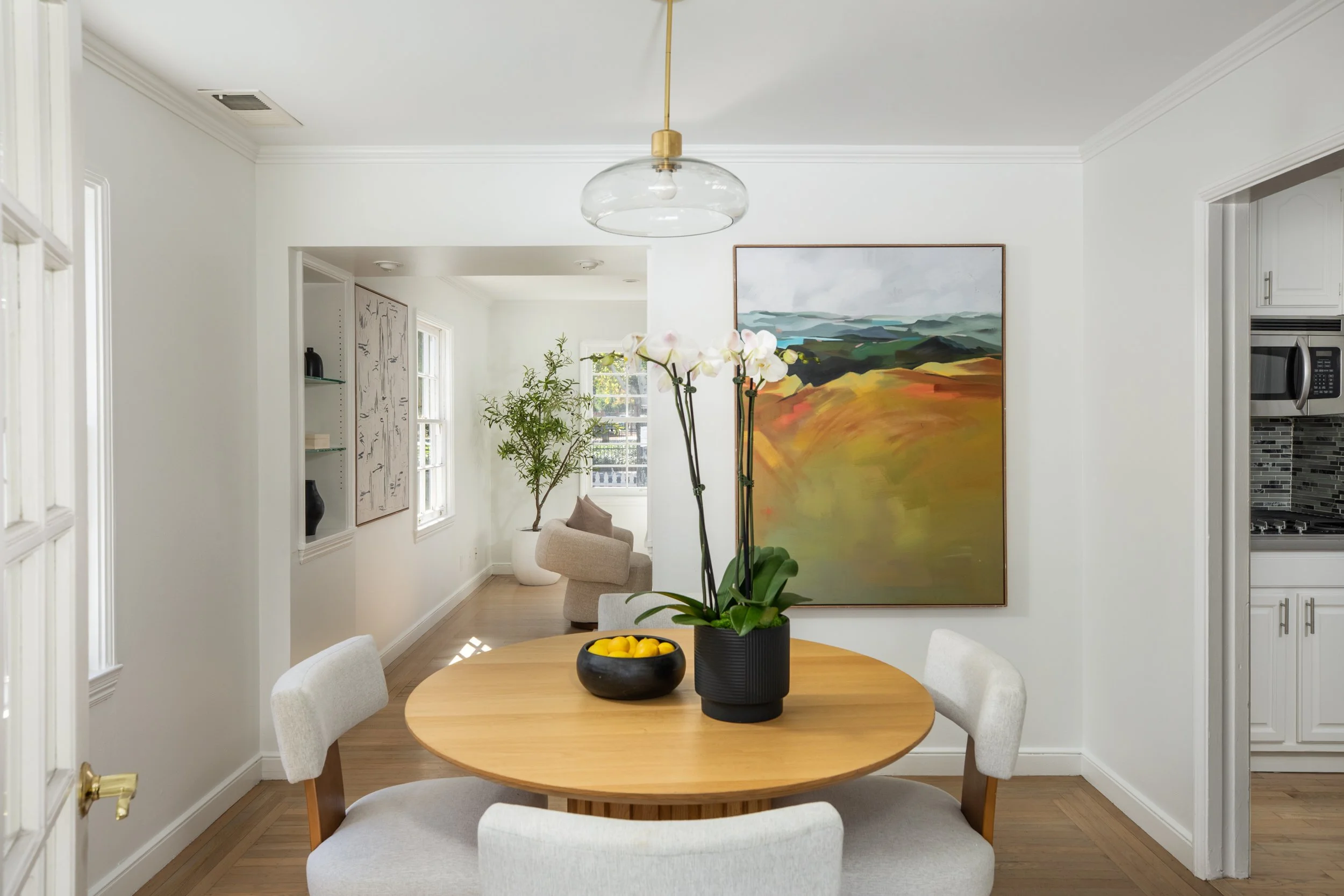 A dining room with a wooden table, four cream-colored chairs, a black vase with white orchids, a bowl of lemons, a large landscape painting, a potted plant, and a modern light fixture hanging from the ceiling.