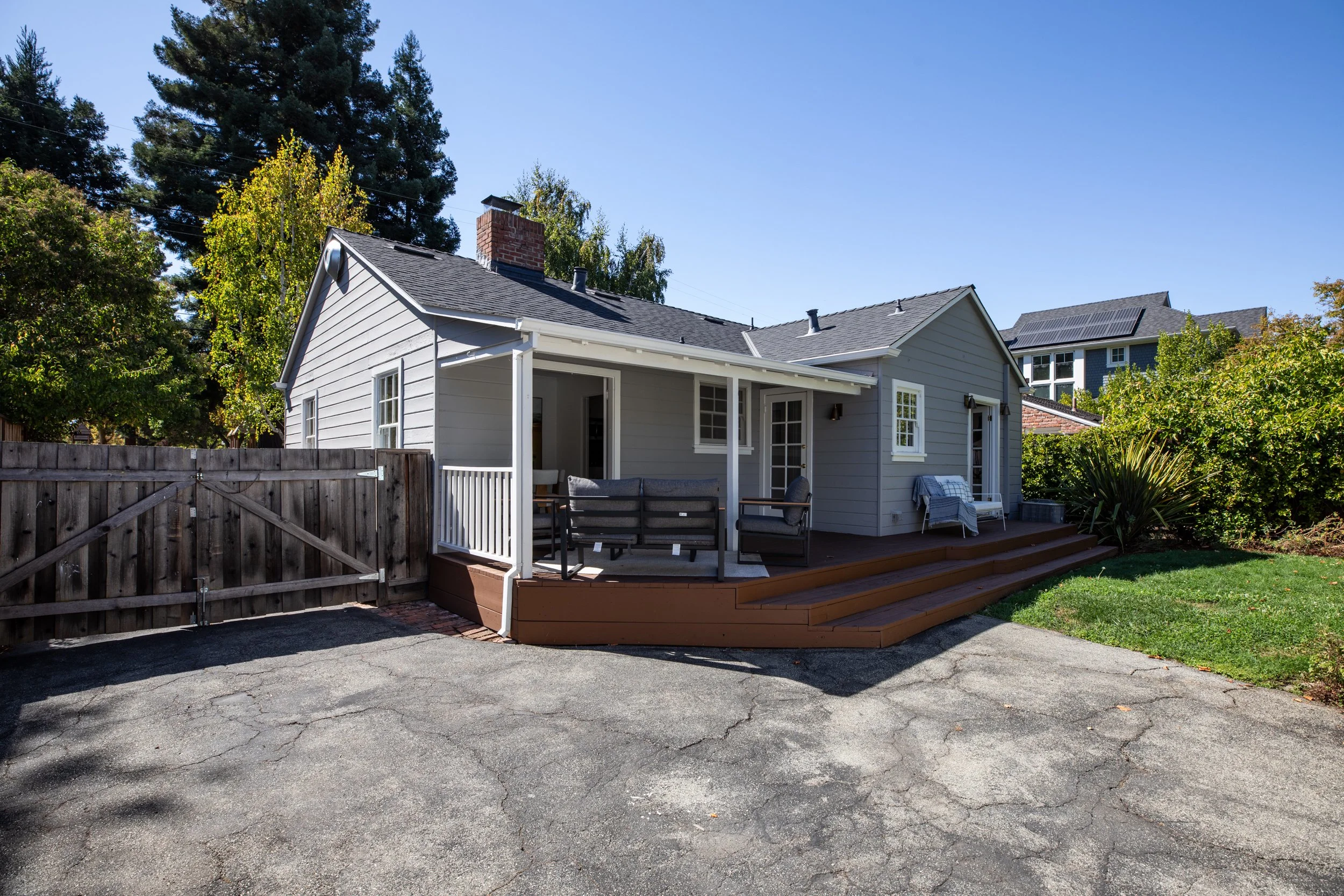 A backyard with a small light gray house, a wooden deck, outdoor seating, and a wooden fence. There is a tree and a clear blue sky.