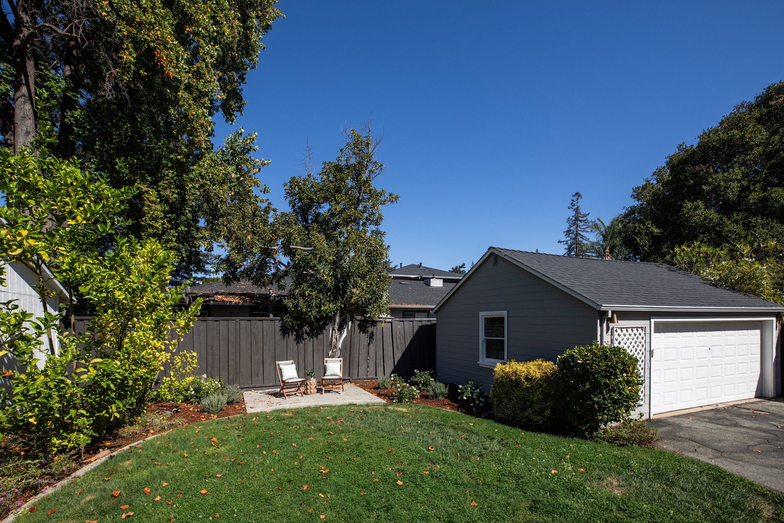 A backyard garden with green lawn, small patio area with two chairs, and a white house with a gray roof surrounded by trees and shrubs. Clear blue sky above.