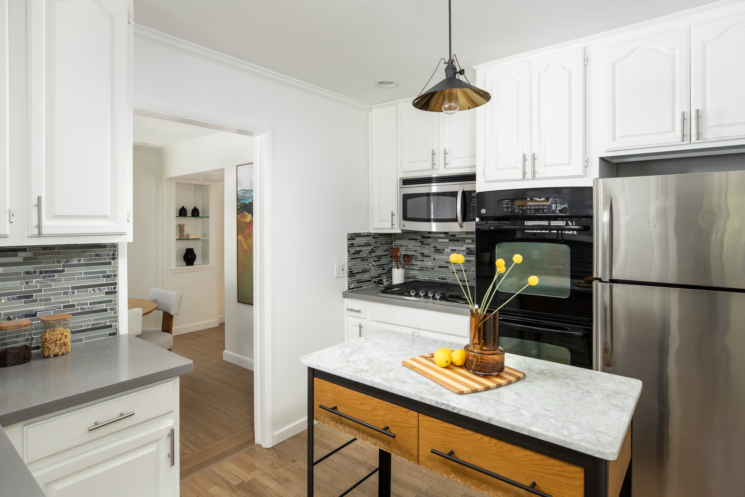 Modern kitchen with white cabinets, black appliances, marble countertops, and a small island with wooden drawers. A vase with yellow flowers on the island, and a backsplash with gray and white tiles. A dining area is visible through a doorway.