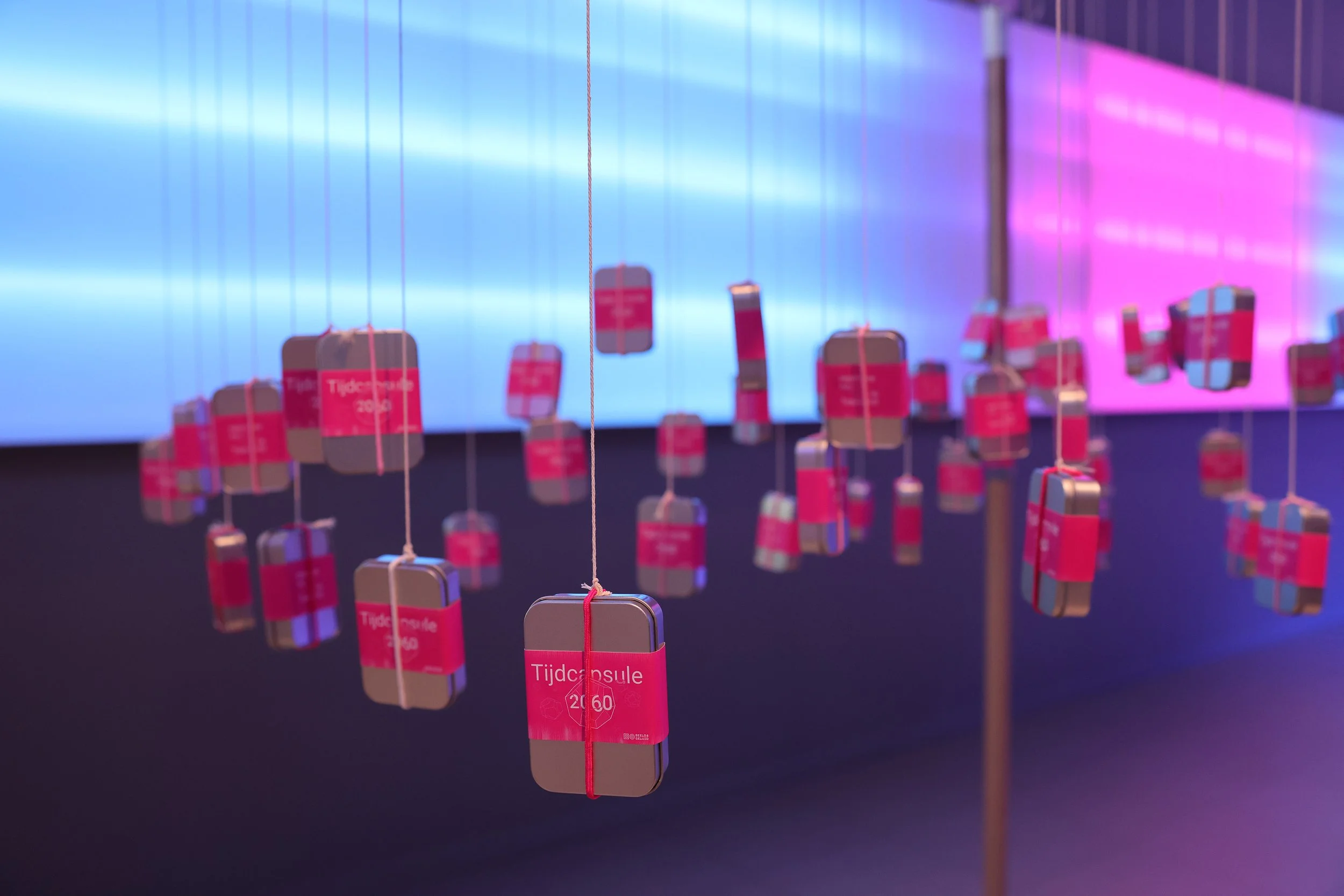 Diminutas placas de metal colgadas por cuerdas, con etiquetas rosas que dicen 'Tijdcapsule' y un año, parece ser una exhibición o una instalación artística con luz de fondo azul y morada.