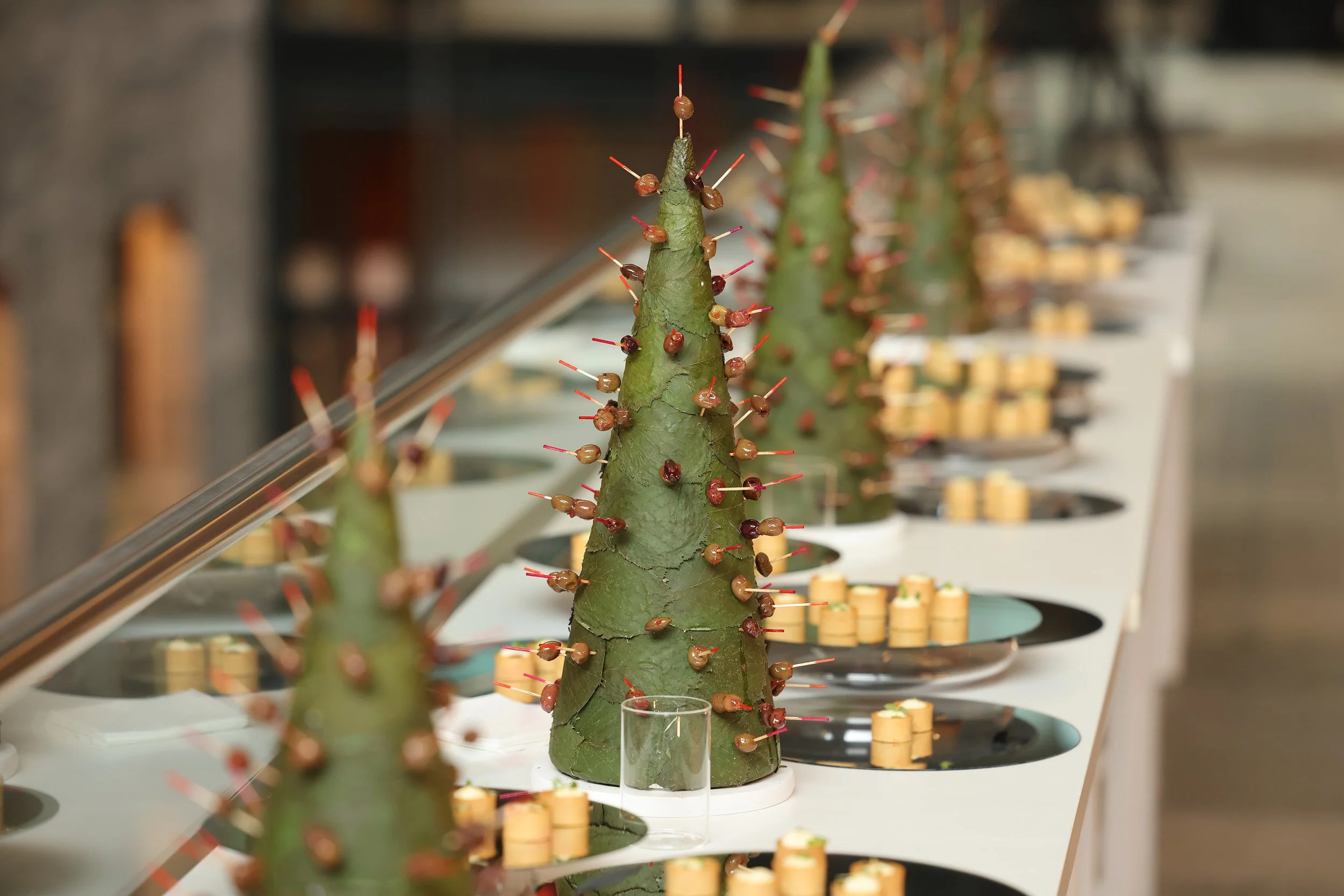 Una piña de cactus decorada con pasas y palillos en un evento o exposición, con varias pequeñas estructuras cilíndricas en color amarillo en el fondo.