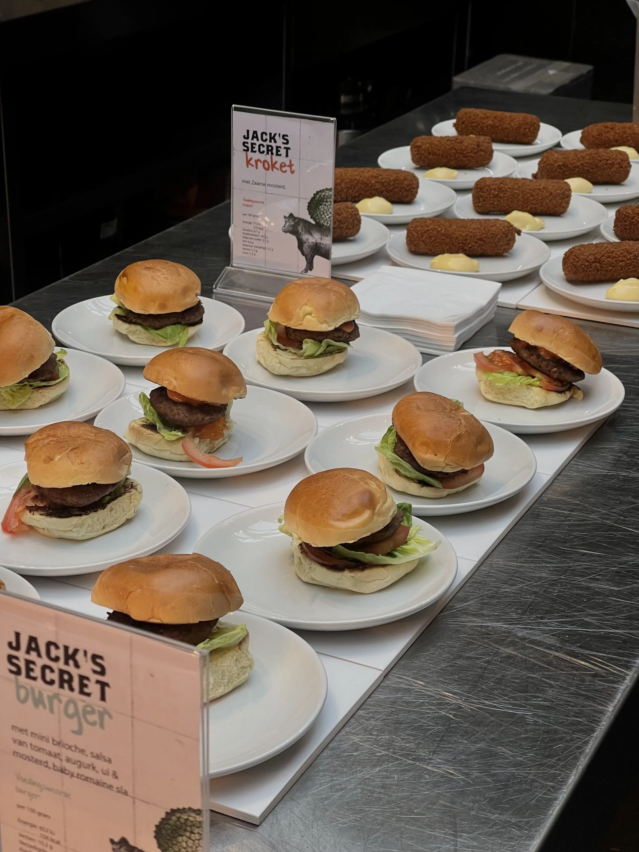 Variedad de mini hamburgesas con pan, lechuga, tomate y carne en platos blancos, y croquetas de pollo con salsa en platos pequeños, en una mesa de buffet.