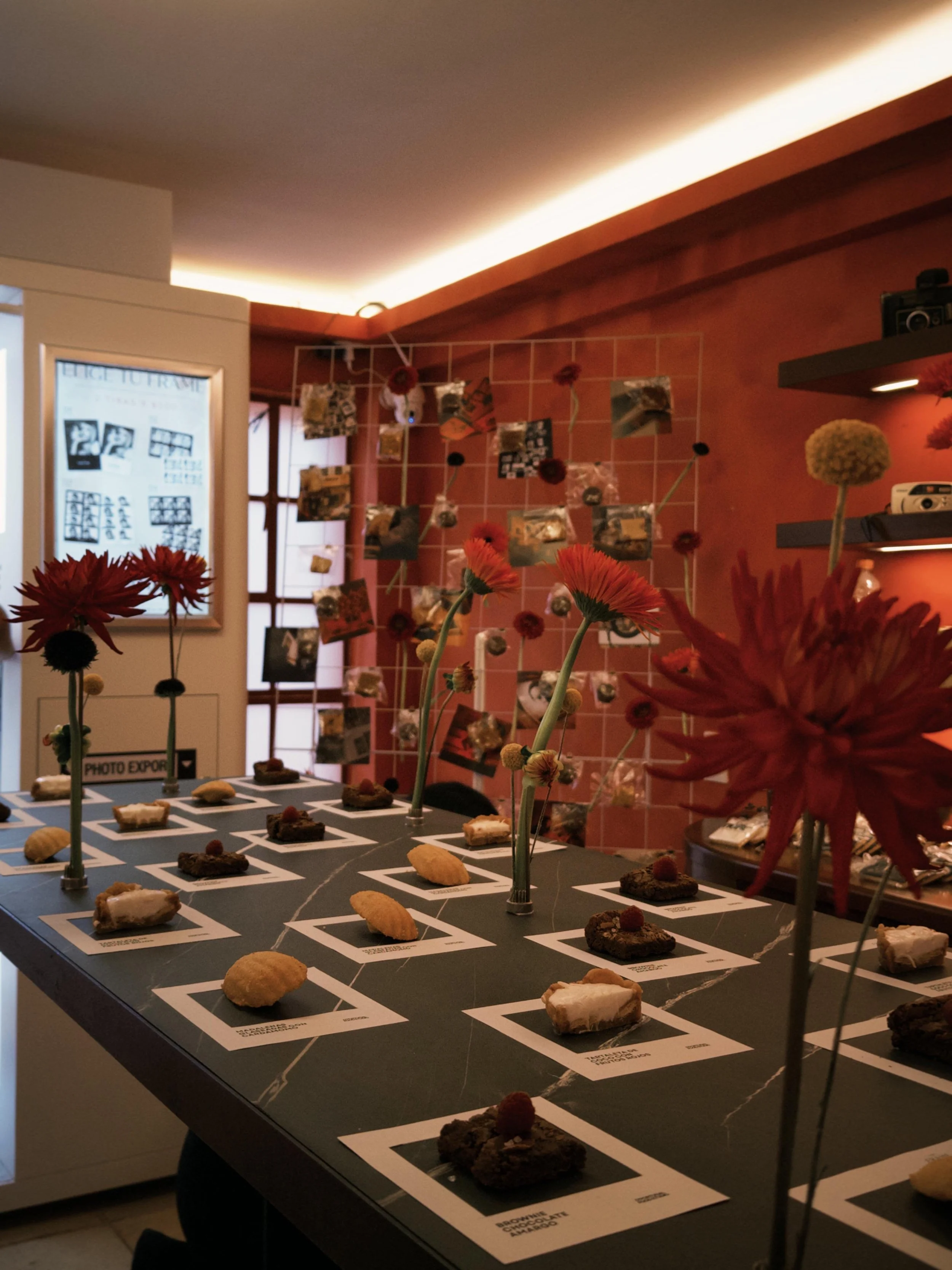 Mesa con varias muestras de postres y dulces, cada uno en su propio espacio con etiquetas. Decoración con flores. Fondo con muro rosa y exhibiciones de fotos colgadas en una estructura.