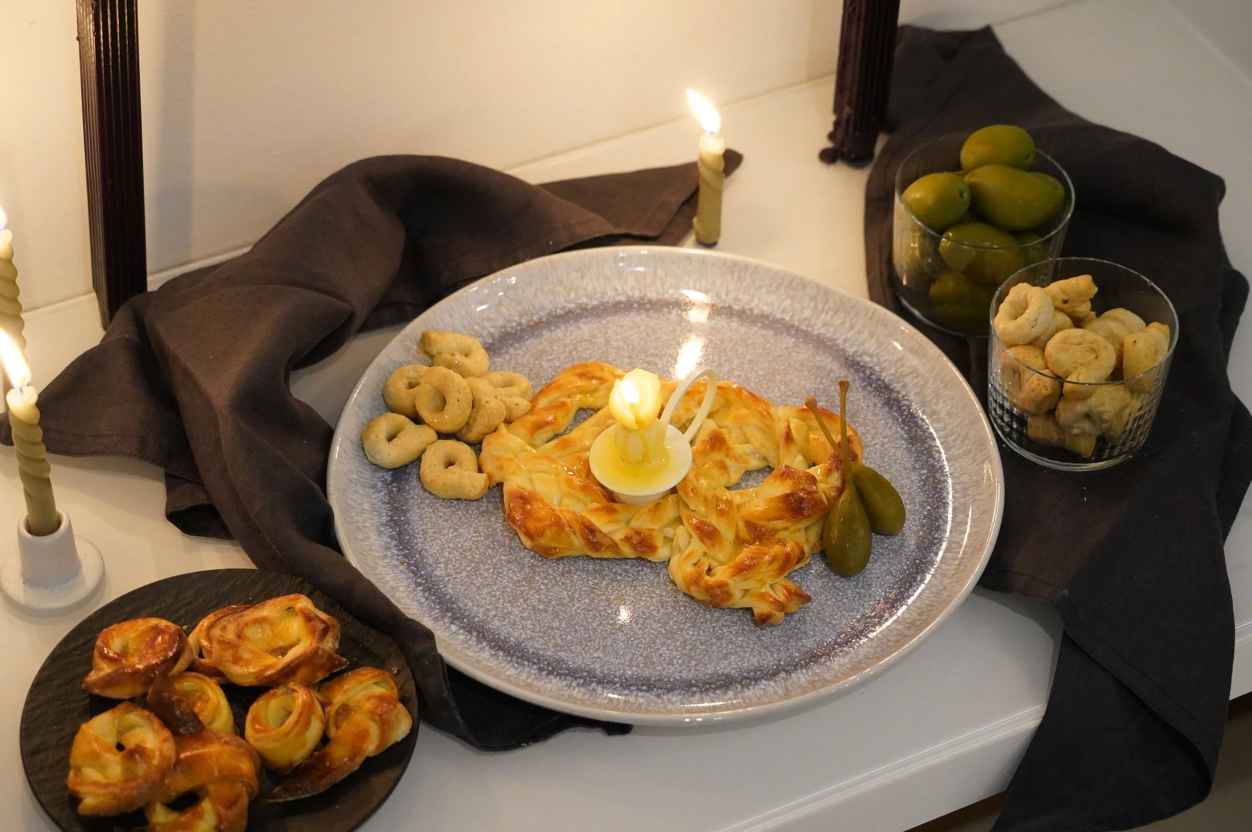 Mesa decorada con comida, una focaccia y varias empanadas pequeñas, acompañado con uvas verdes y galletas saladas en vasos de vidrio. Con una vela de mantequilla al centro