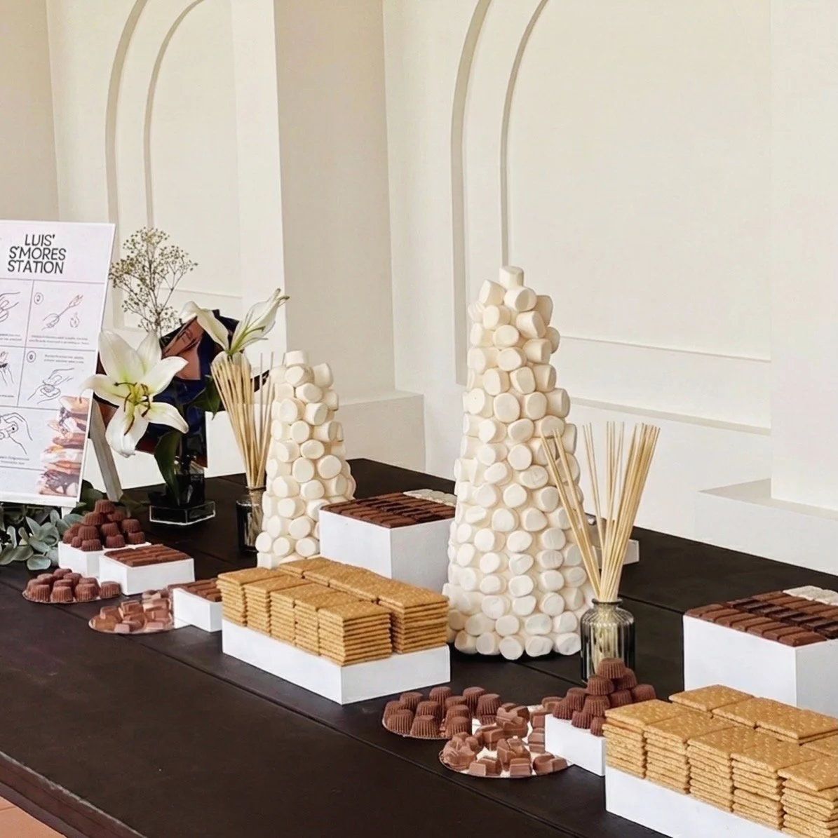 Mesa decorada con varias torres de malvaviscos, galletas y chocolates, con jarras con palitos de madera y flores blancas en un jarrón.