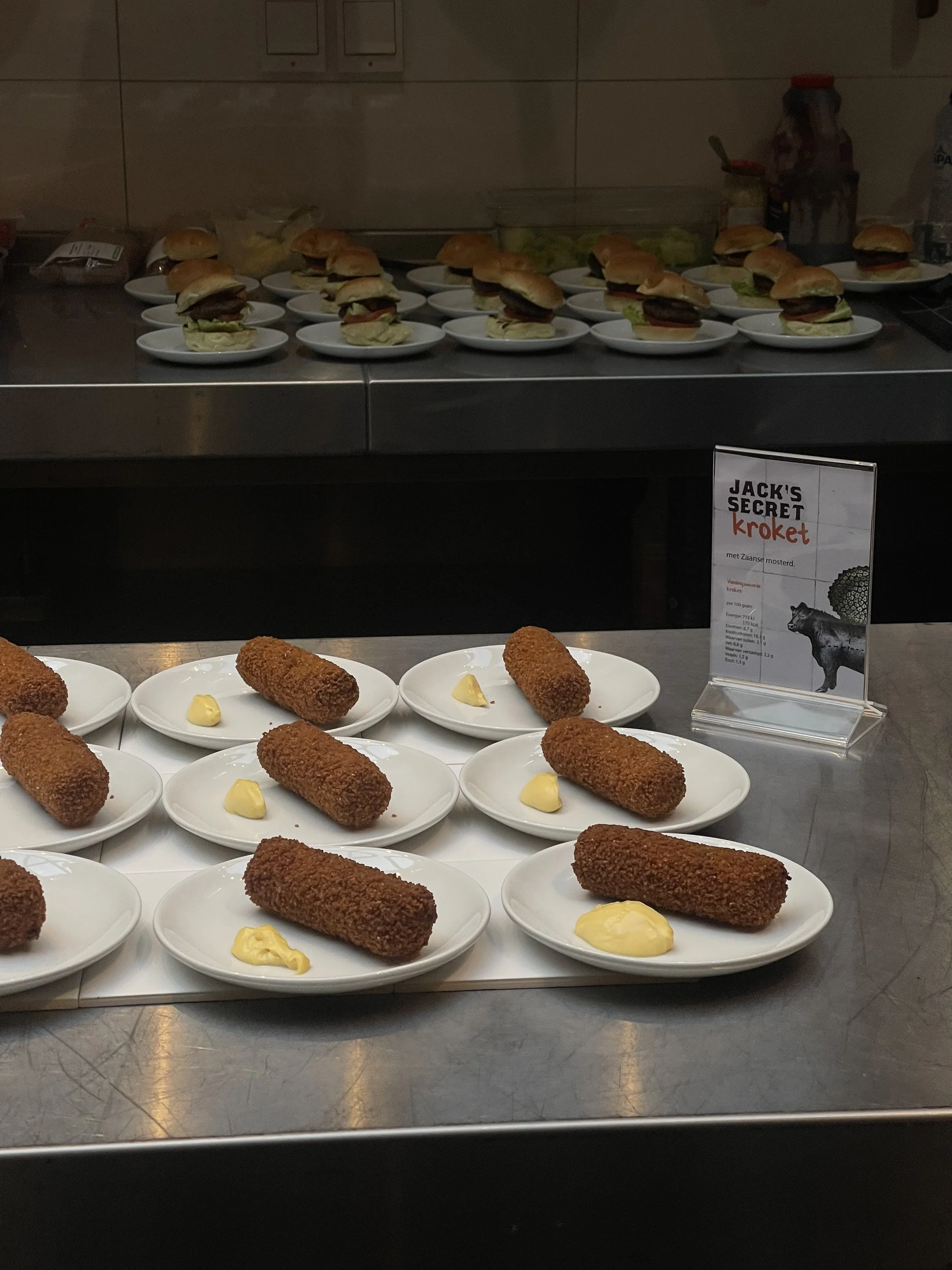 Varios sándwiches pequeños con carne, lechuga, tomate en panes suaves, colocados en platos blancos. En primer plano, hay croquetas de tamaño mediano acompañadas con una cucharada de mantequilla o salsa en platos blancos. En el fondo, una señal promocional en un cartel de plástico ofrece información sobre una croqueta con mostaza de Zaanse. La escena es de una exposición o muestra de alimentos en una superficie metálica.