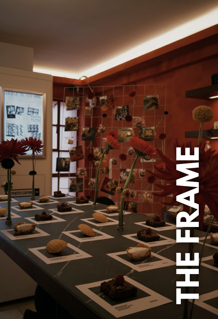 Muestra de varios postres en un exhibidor, decorada con flores y fotos en la pared de fondo en un espacio interior con iluminación ambiental cálida.