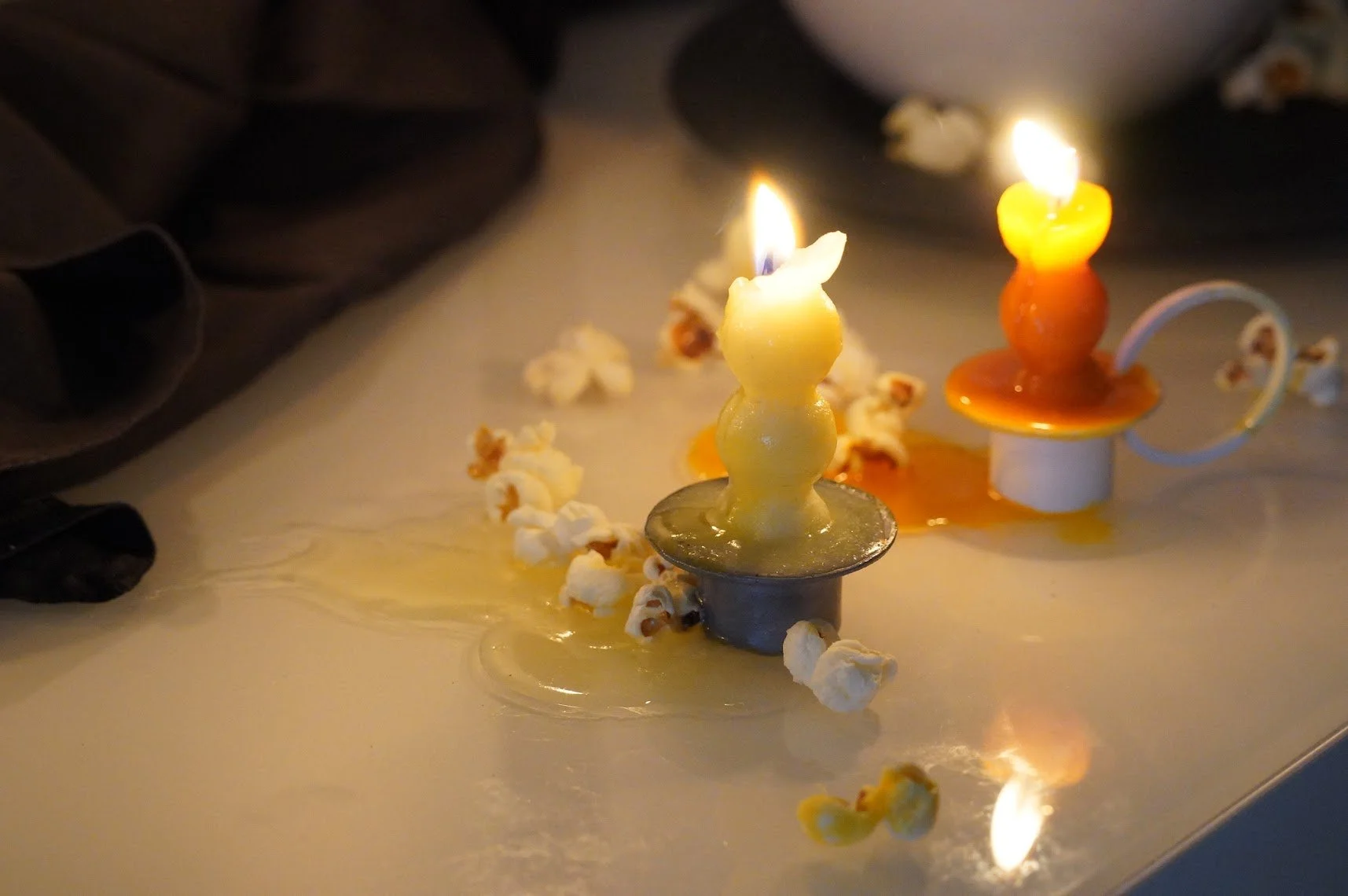 Velas de mantequilla encendidas sobre una mesa, rodeadas de palomitas de maíz y una mano con guantes negros en el lado izquierdo.