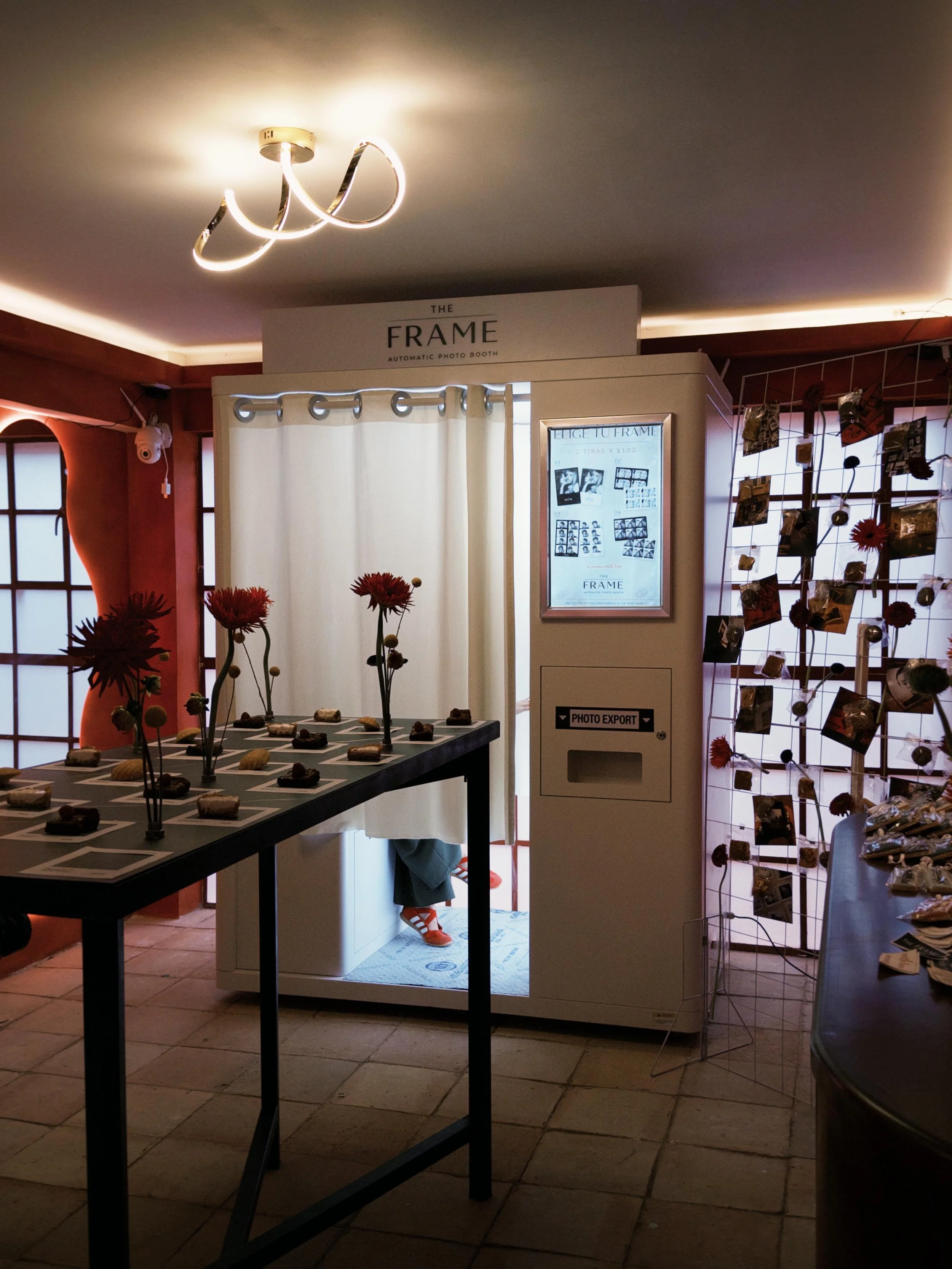 Interior de un estudio de fotografía con una cabina de fotos automática, un cartel que dice 'The Frame', y una mesa con algunos objetos decorativos, junto a una pared con detalles en color coral y una estructura metálica con fotos colgadas.